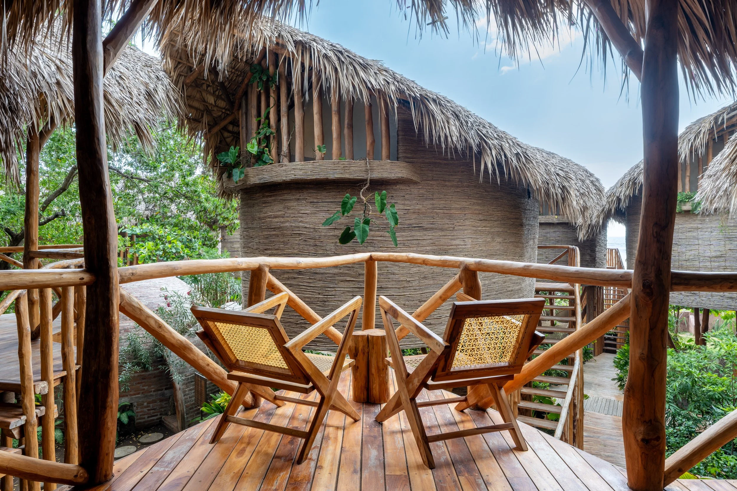 Wooden balcony with chairs overlooking huts with thatched roofs, surrounded by green trees under a blue sky.