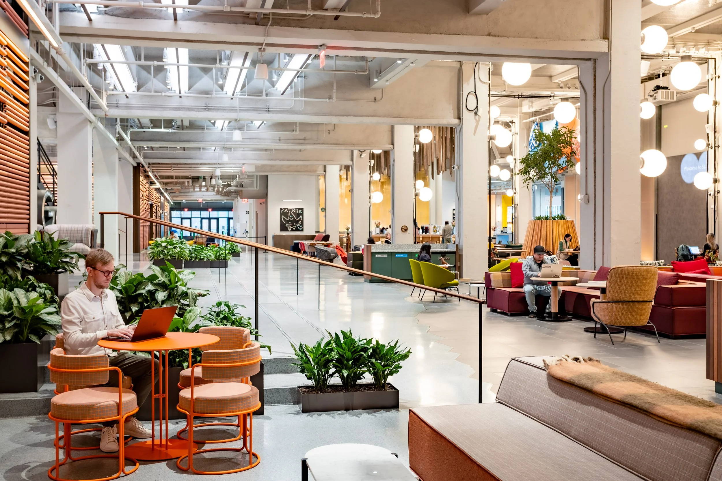 Interior of a modern, well-lit public space or lobby with seating areas, potted plants, and people using laptops and devices.