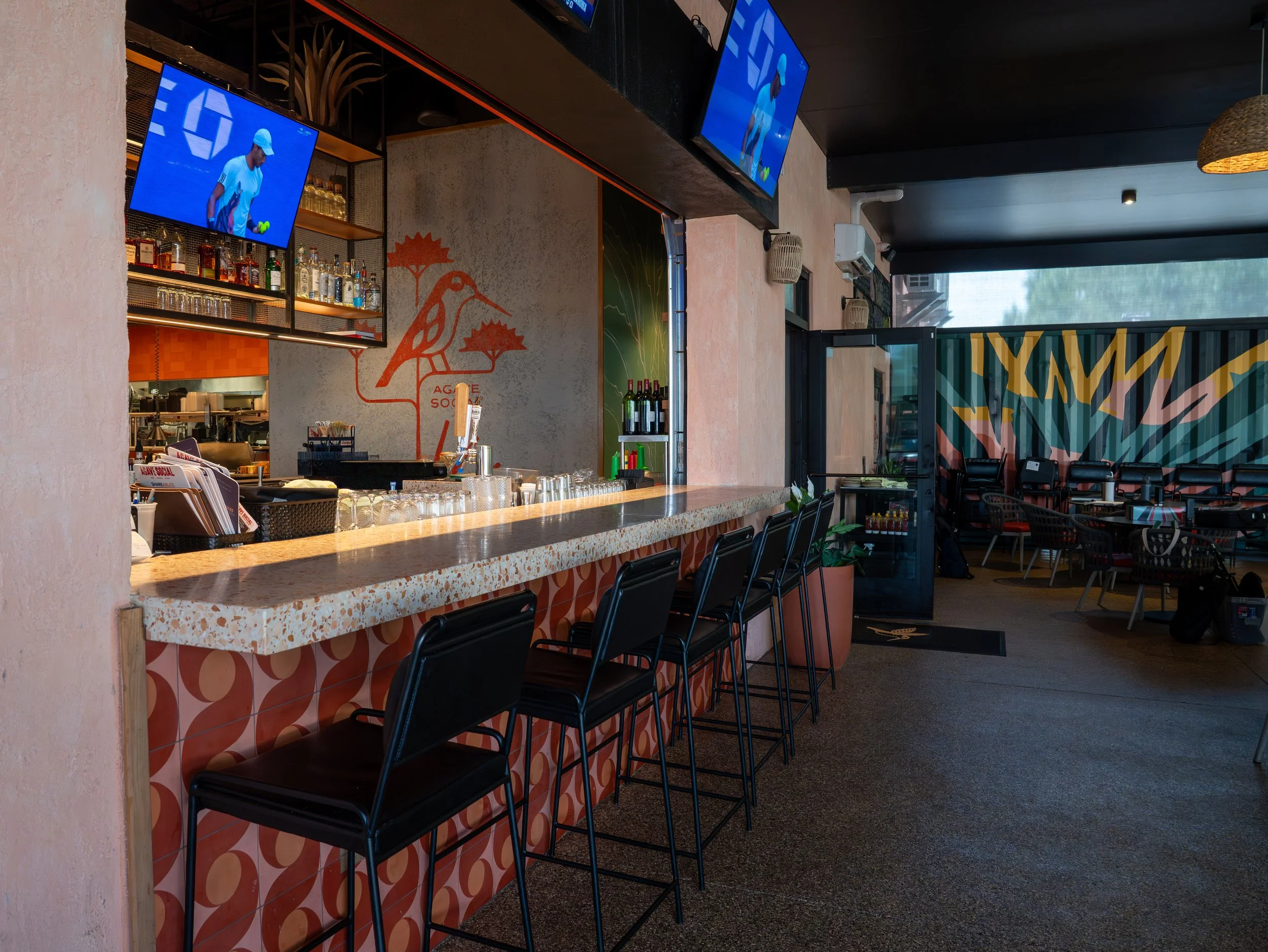 Interior of a sports bar with a terrazzo bar counter, black chairs, and multiple television screens displaying a tennis match. There is a colorful mural on the wall and a seating area in the background.