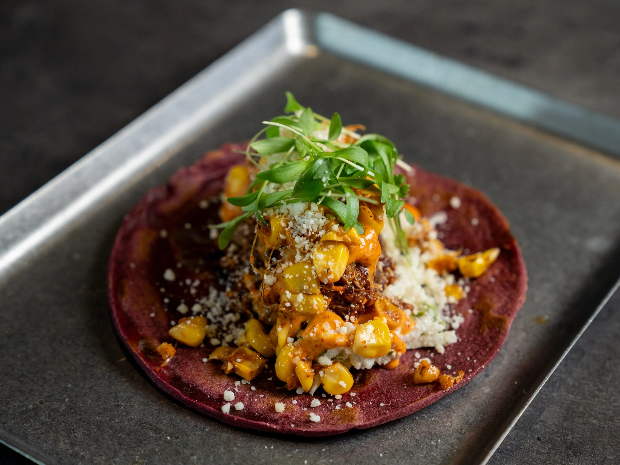 Close-up of a gourmet dish with a purple tortilla, topped with corn, cheese, and microgreens, served on a metal tray.