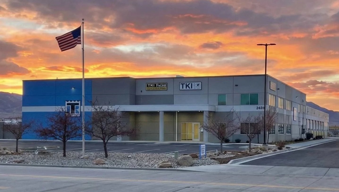 A commercial building with a blue and gray exterior and signs that read 'TKI Freight' and 'TKI' at sunset, with an American flag on a flagpole in the foreground, planted trees, and mountains in the background.