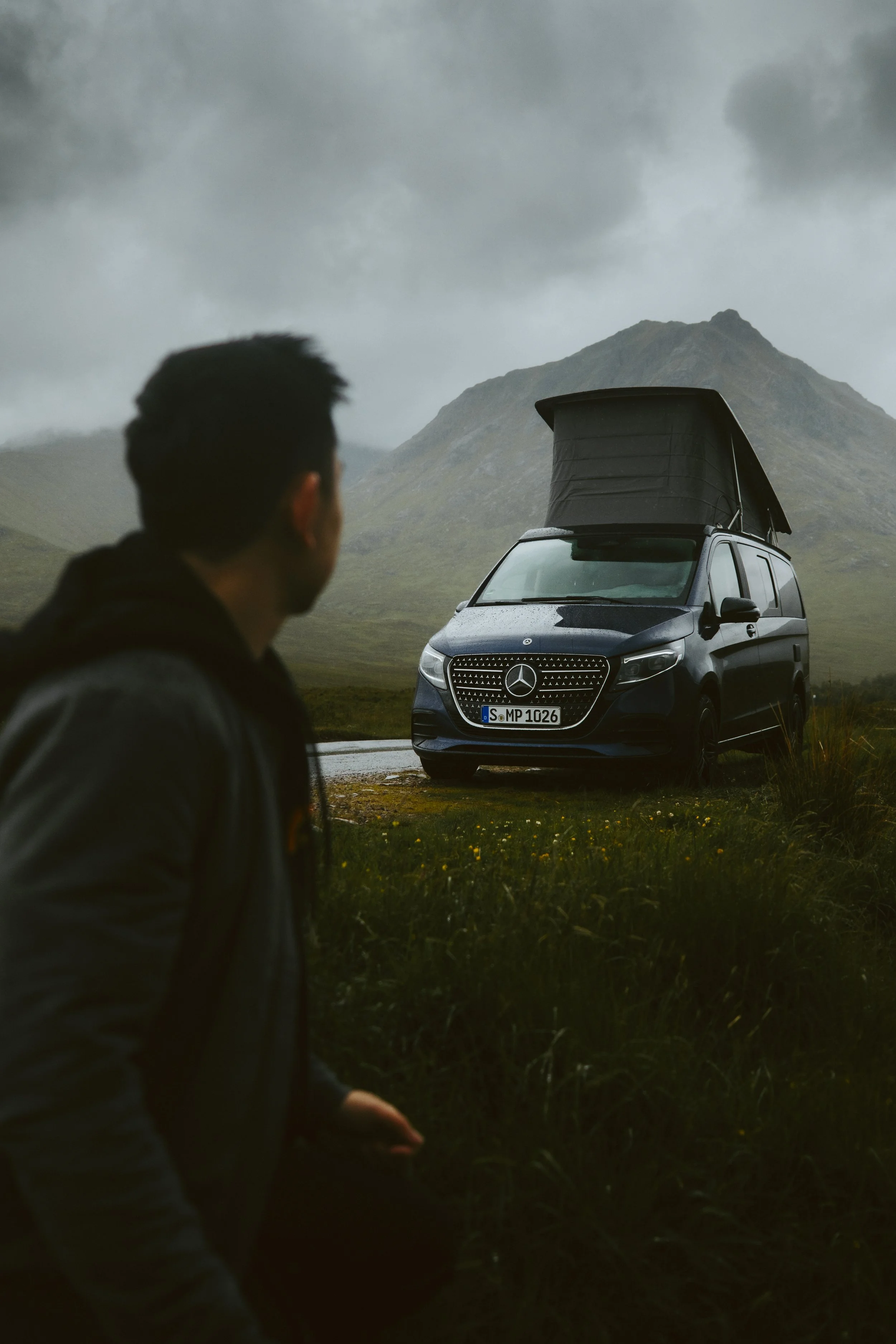 A person with a backpack looking at a black camper van with a pop-up roof parked on a grassy area in a mountainous landscape on a cloudy day.