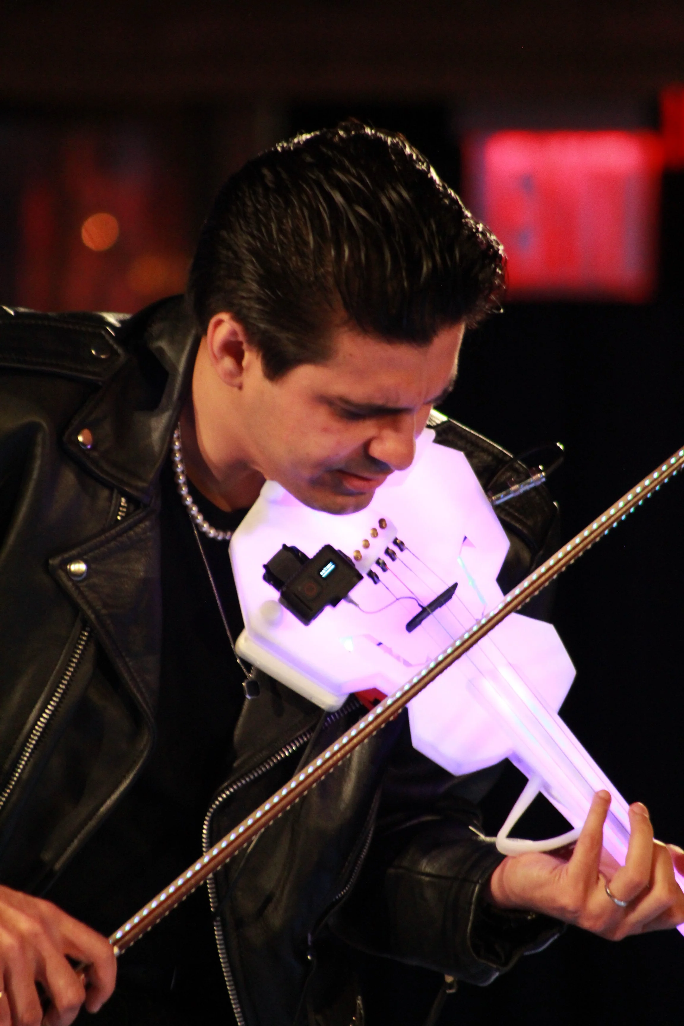 A man with black hair in a leather jacket playing a glowing, sci-fi style white electric violin at night.