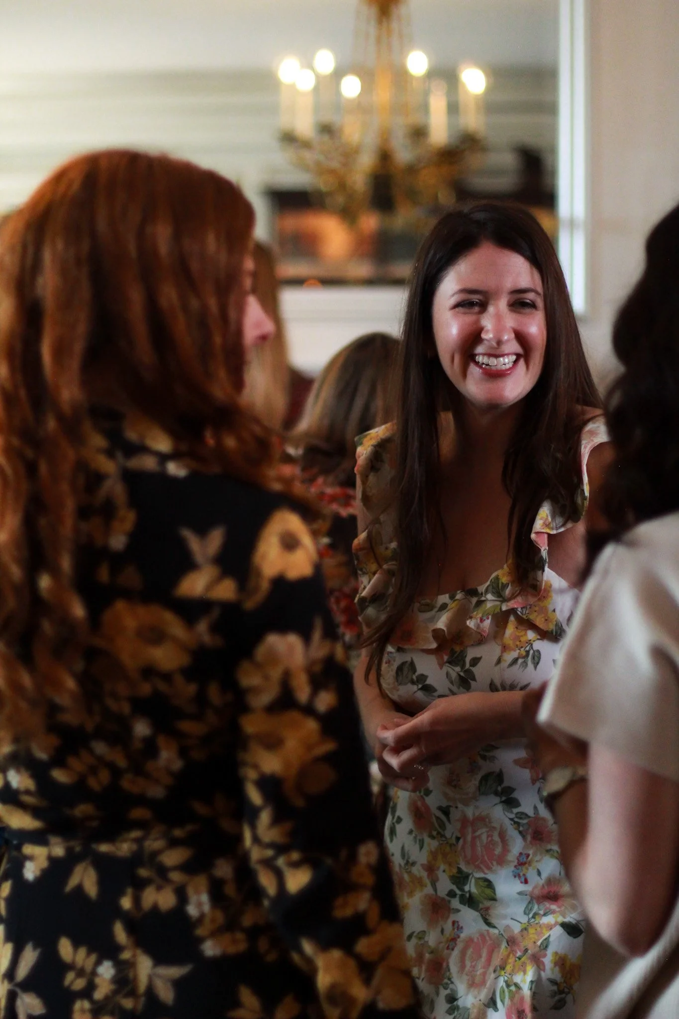 A woman with long dark hair and a floral dress smiling and talking with others in a social gathering indoors, with a chandelier in the background.