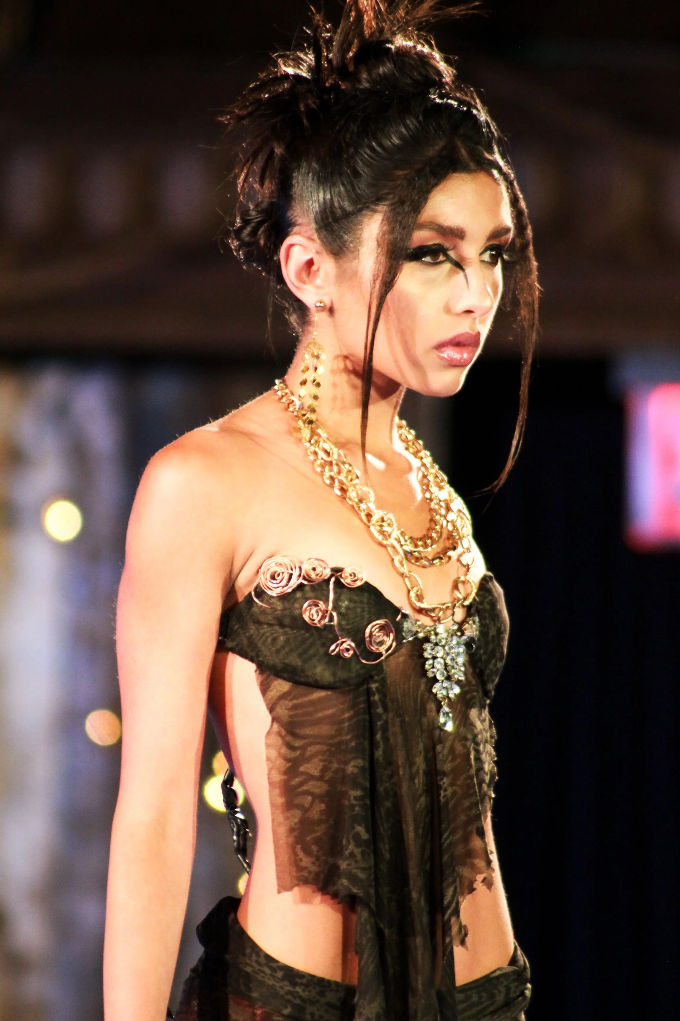 Female model with dark hair styled in an updo, wearing large gold jewelry, a black sheer top with pink floral detail, and dark pants, walking on a runway.
