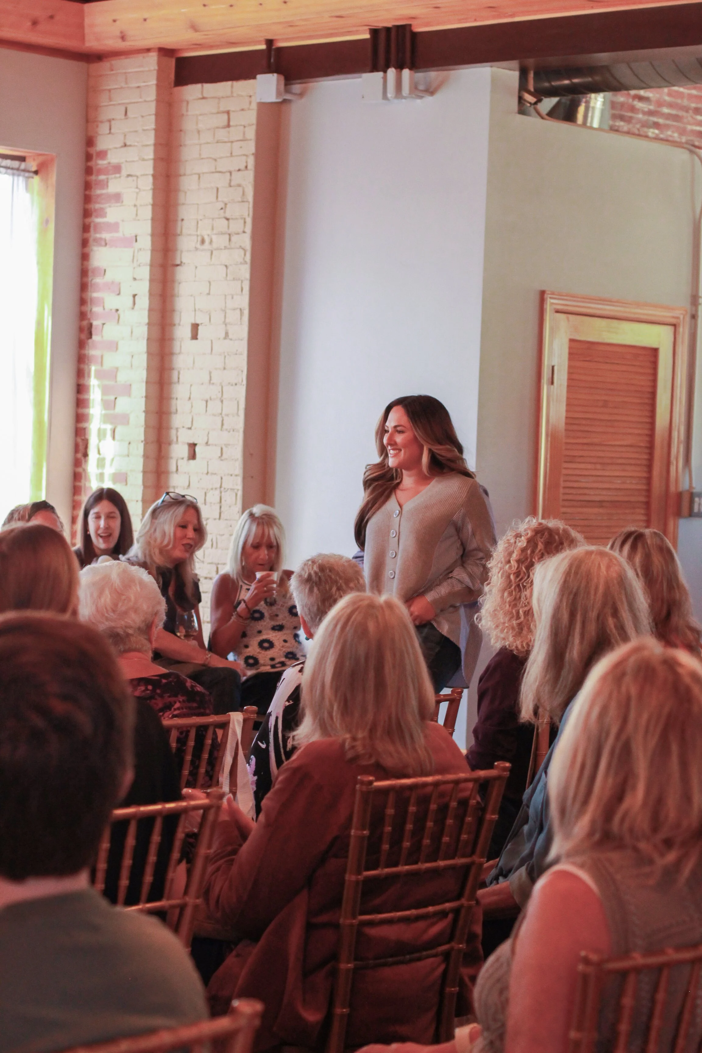 A woman stands and speaks to a seated audience in a cozy, warmly lit room with exposed brick and wood accents.