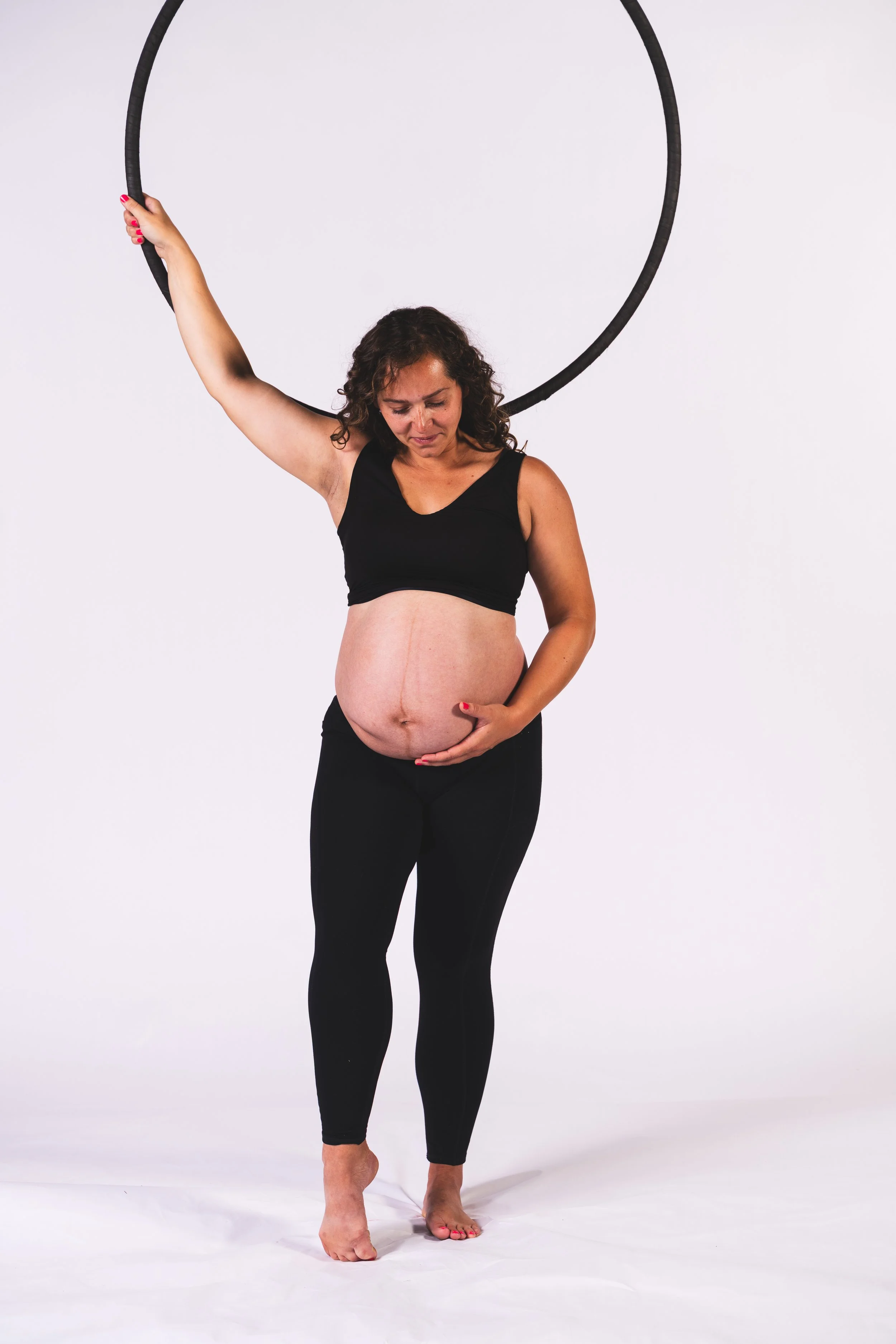 Pregnant woman with curly hair wearing black sportswear holding a large black hoop overhead and gently resting her other hand on her pregnant belly against a plain white background.