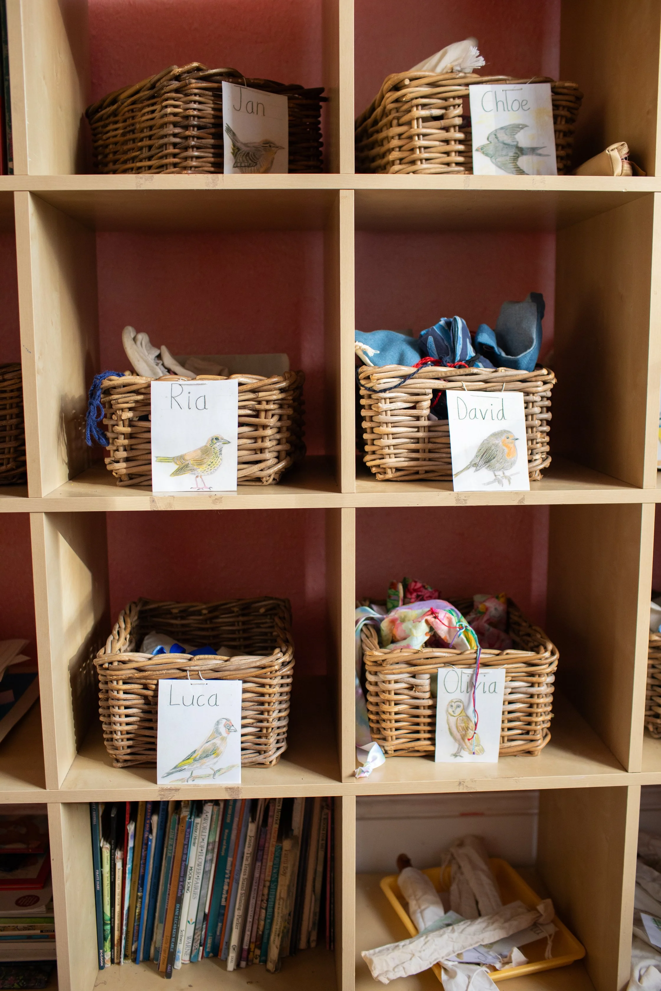 Six wicker baskets on wooden shelf with labeled cards featuring watercolor bird illustrations: Jan, Chloe, Ria, David, Luca, Olivia.