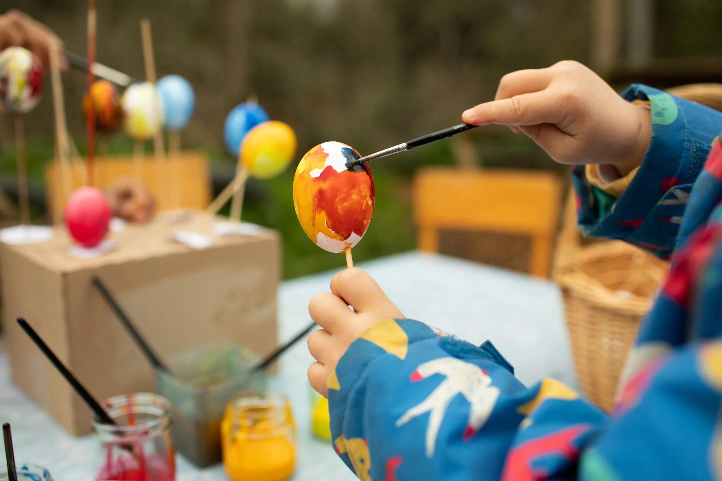 Child painting a decorated Easter egg outdoors with jars of paint nearby and other colorful eggs hanging in the background.