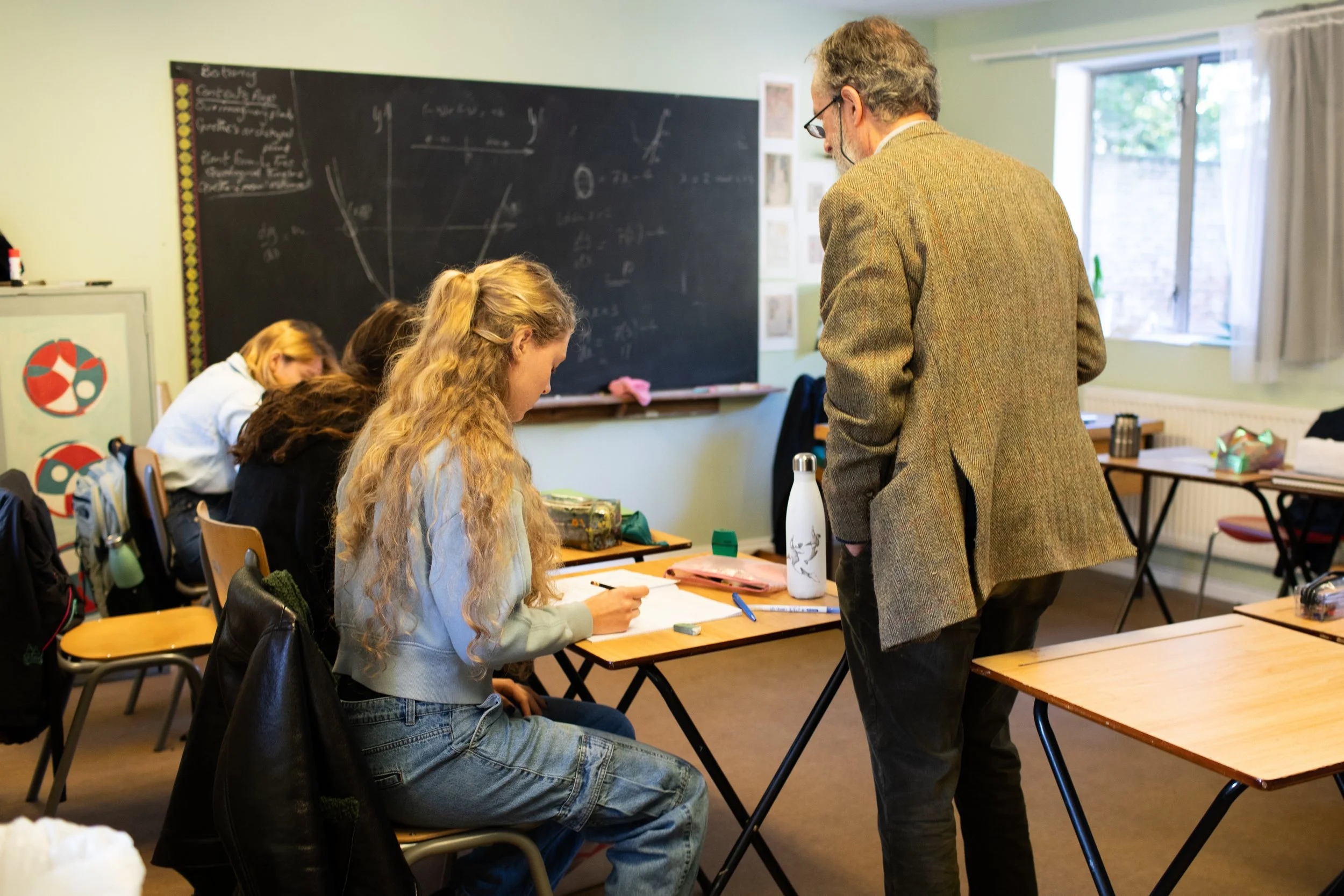 A classroom scene with a teacher standing next to a seated female student, with other students in the background near a blackboard.