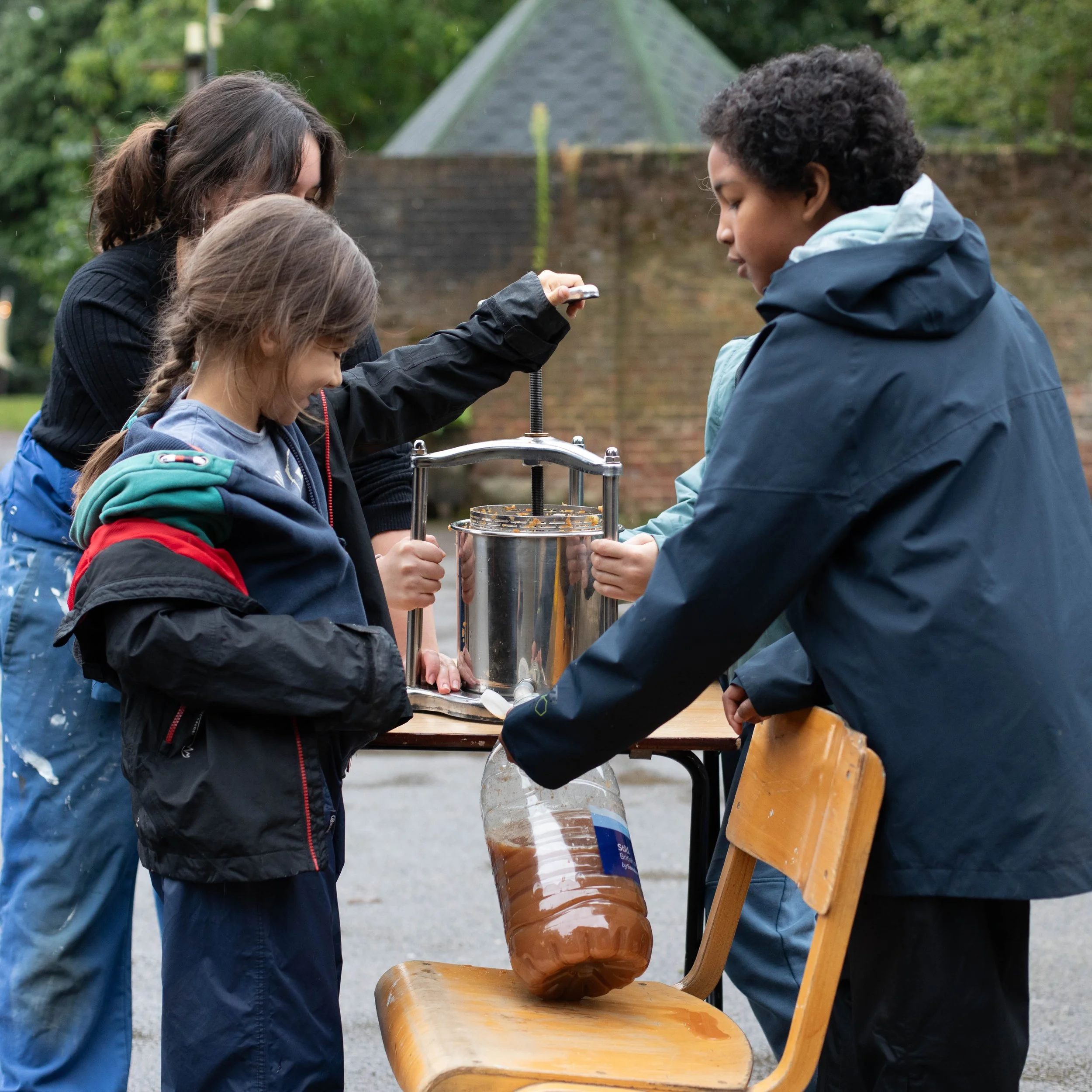 A group of four children pouring and pressing orange juice using a manual juicer outdoors during daytime.