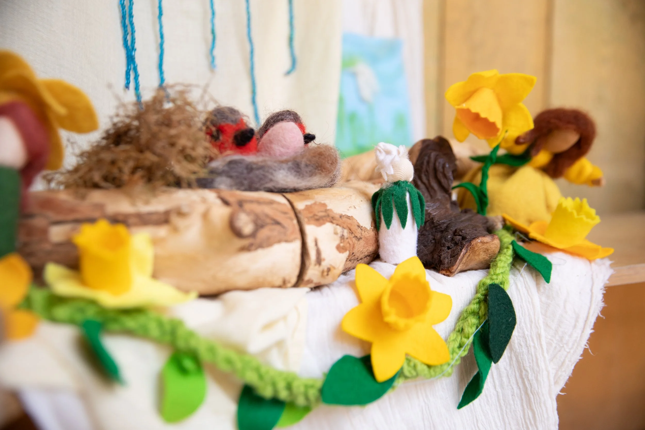 Felted animal and flower figures arranged on a wooden surface, including moss, logs, and paper flowers.