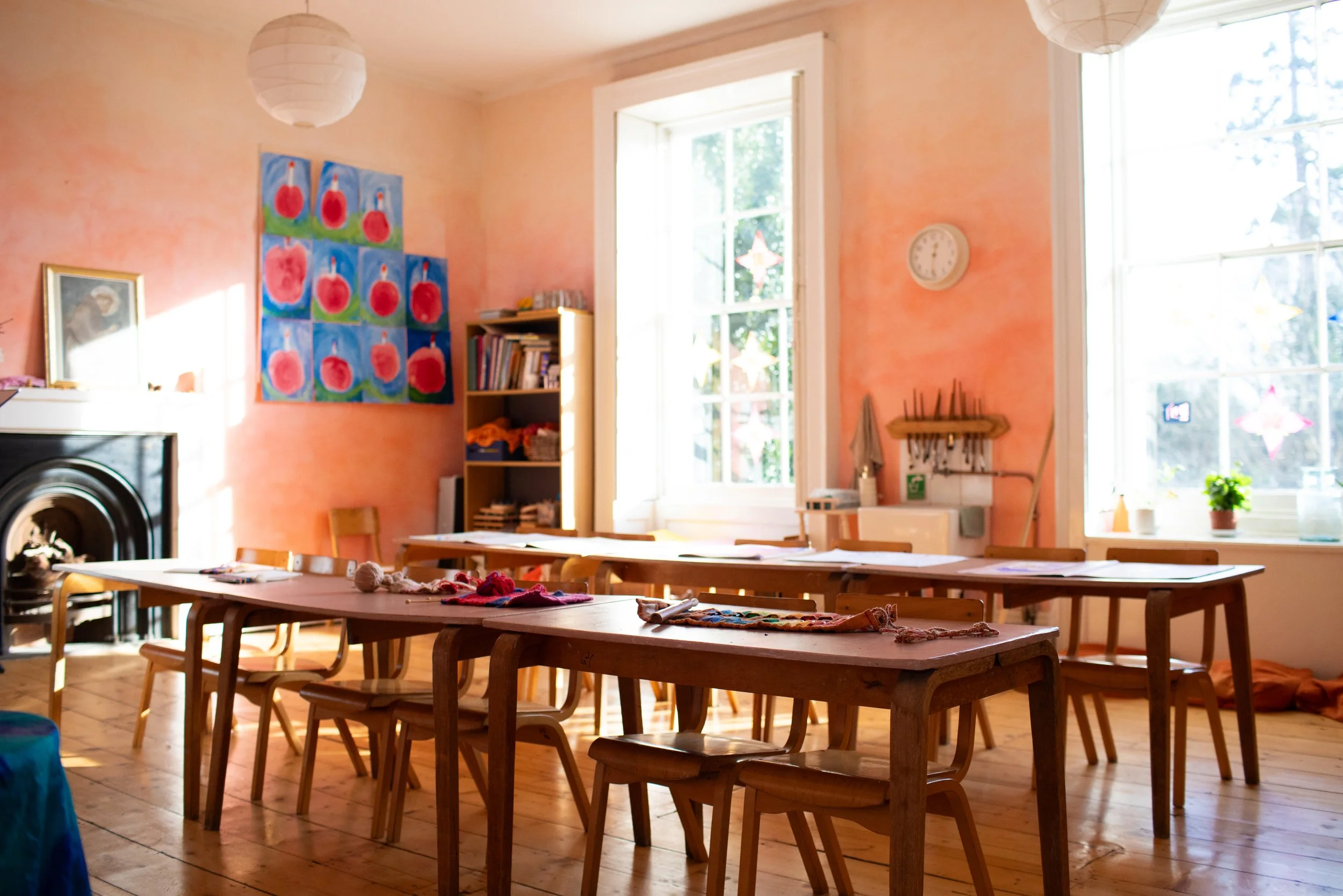 Bright classroom with pink walls and large windows, wooden tables and chairs, colorful artwork on the wall, bookshelf with books, and craft materials on the tables.