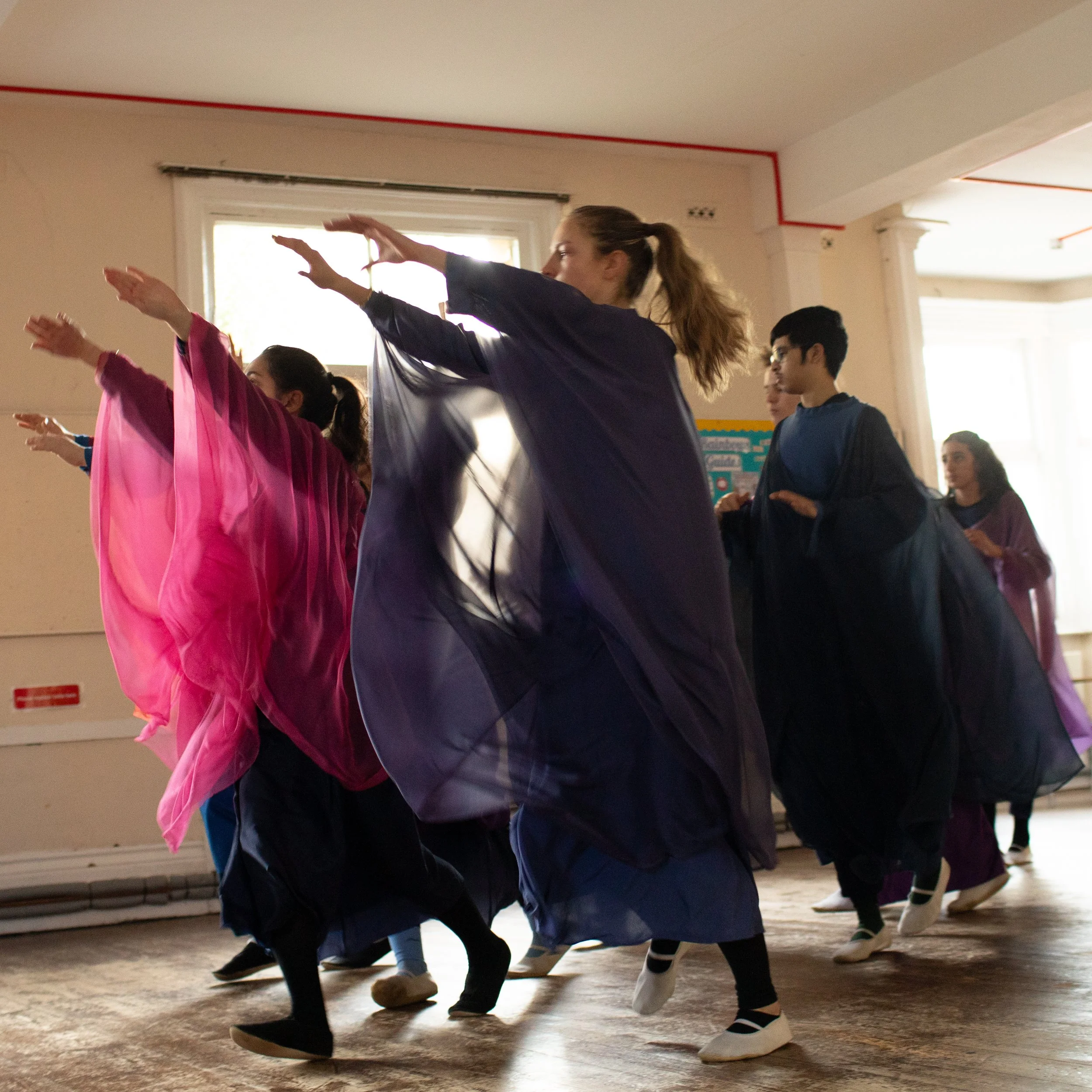 Group of young people dancing in a room with large windows and wooden floors, dressed in colorful traditional dresses.