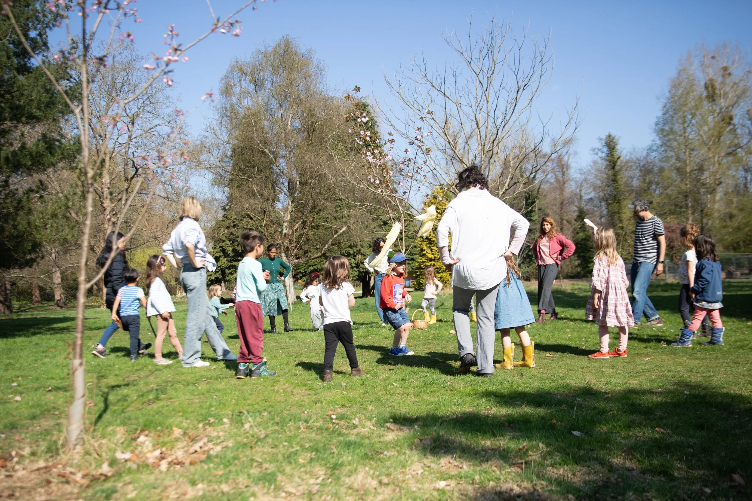 Children and adults gathered outdoors on a sunny day, participating in a game or activity in a park with trees and grass.