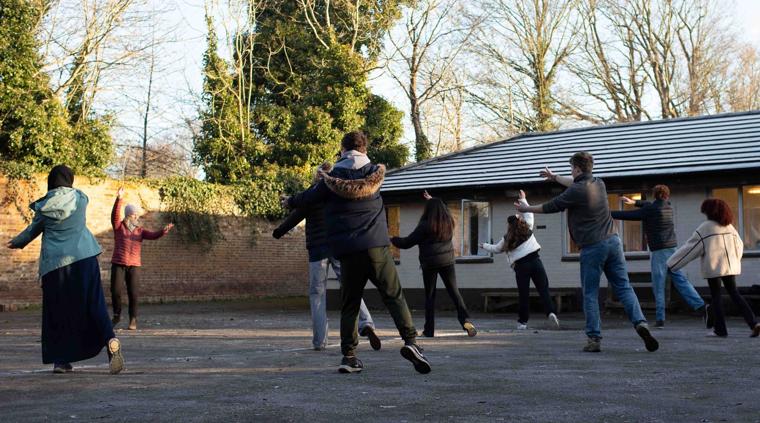 People dancing outdoors in a courtyard with trees, a brick wall, and a building with a metal roof.