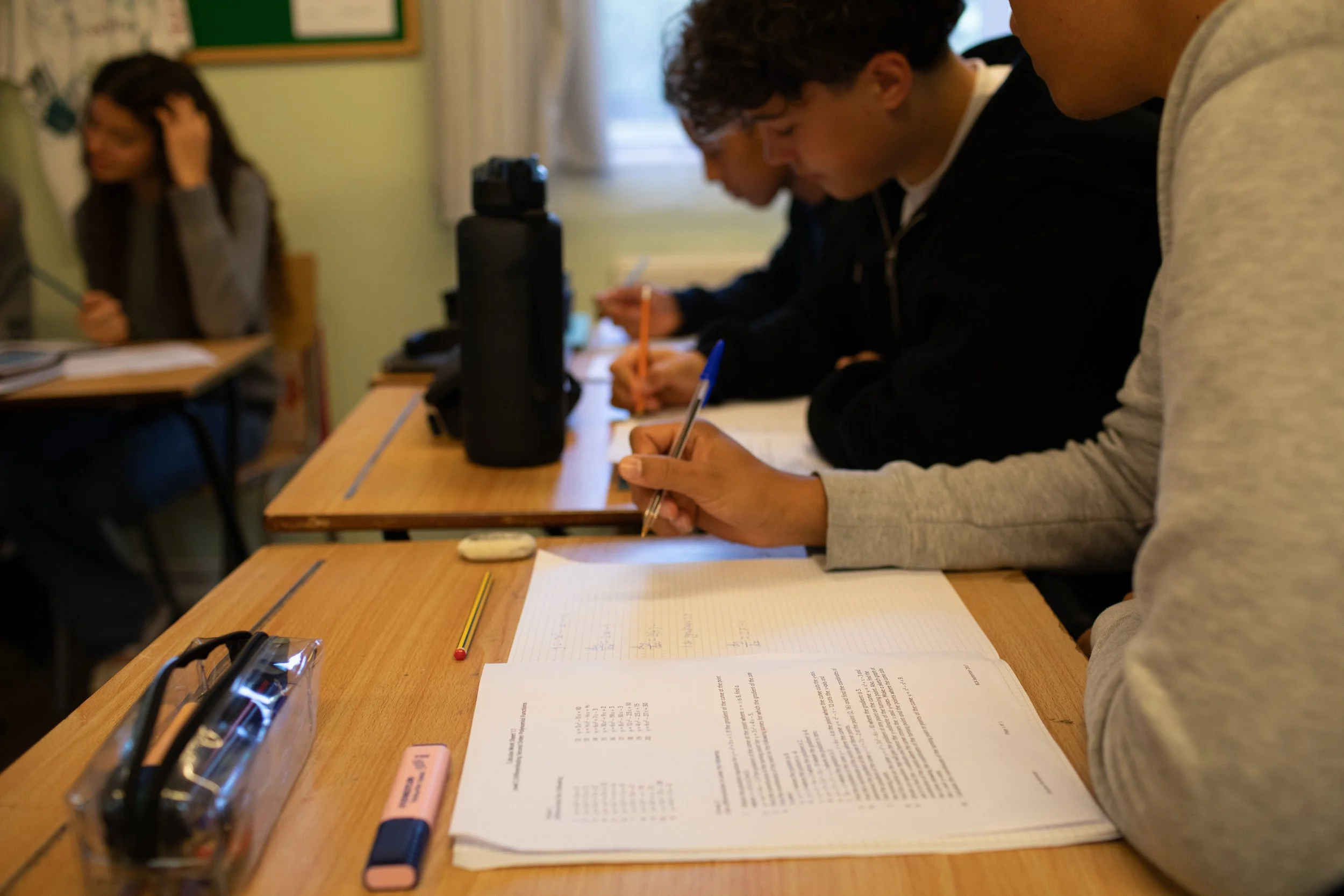 Students sitting at desks in a classroom, taking notes and studying with papers, pens, and backpacks on the desks.