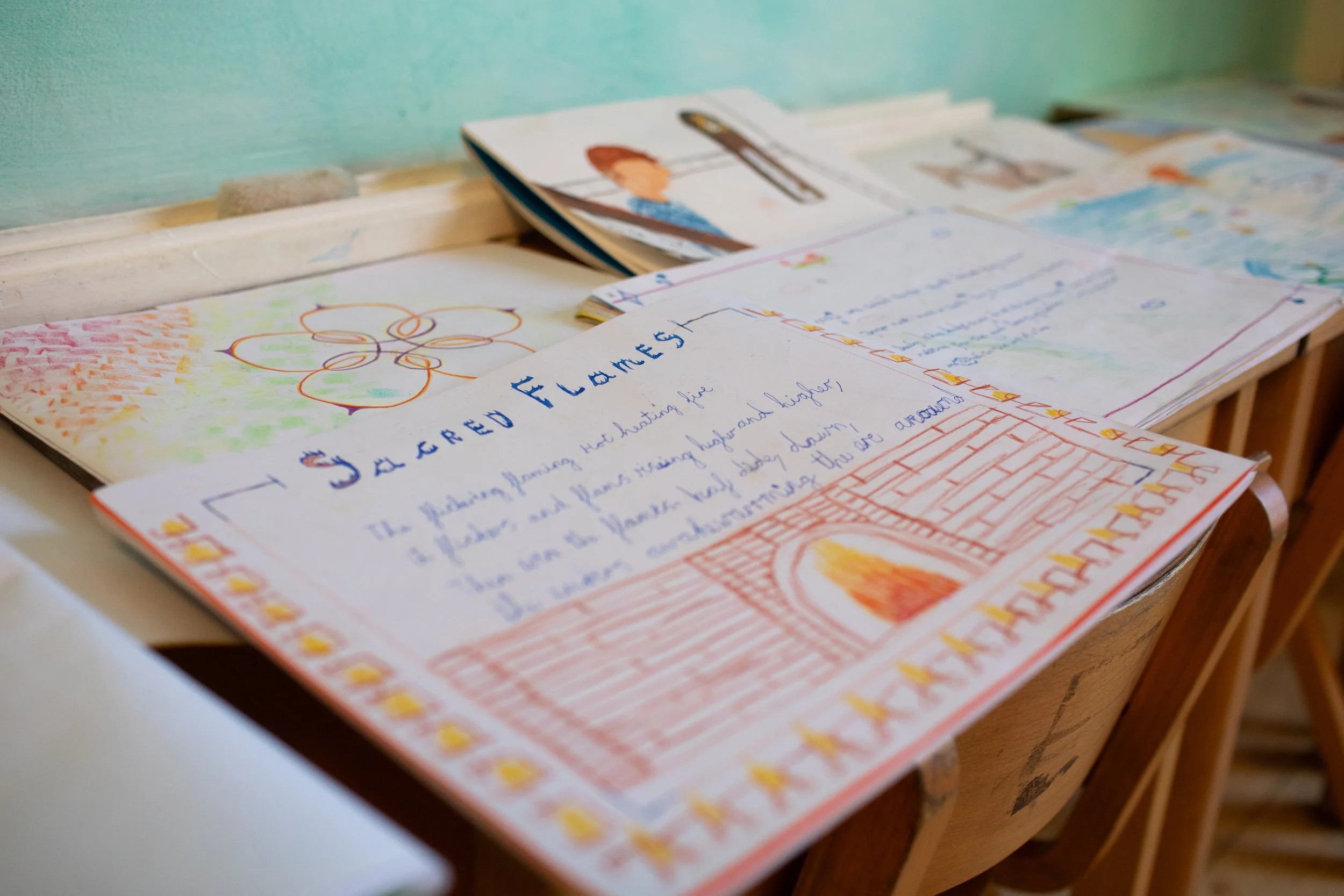 Student project with decorated pages, including a drawing of a fireplace and colorful designs, placed on a classroom desk.