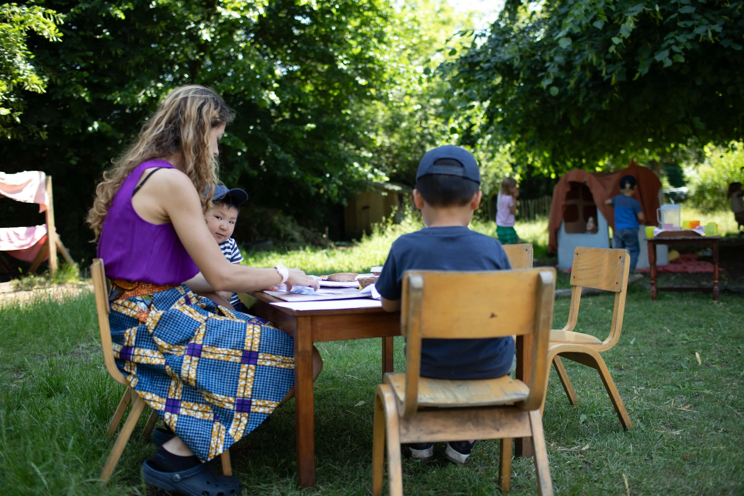 A woman and children sitting at a wooden table outdoors in a lush, green backyard. Two children are seen in the background near a small tent and play area.