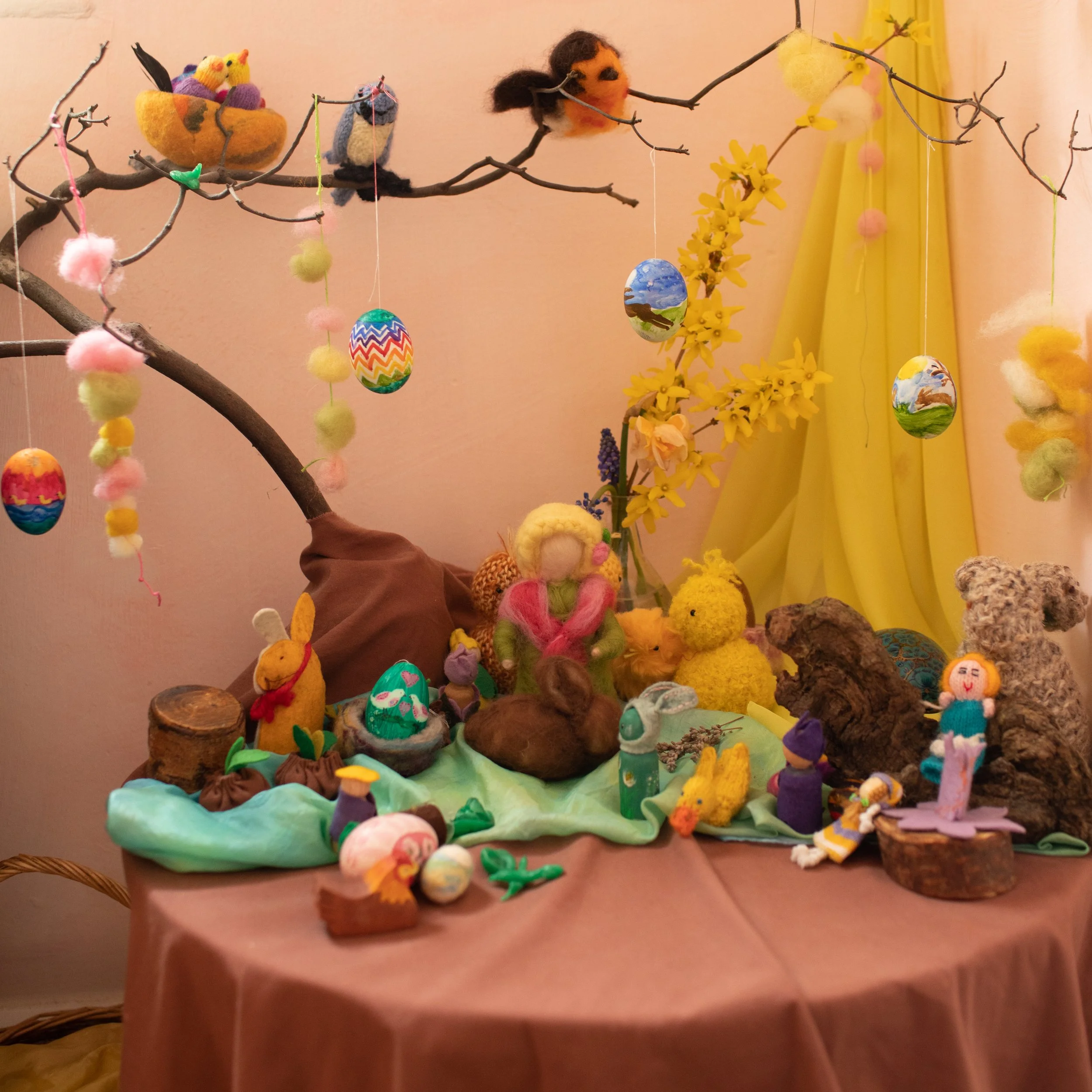 Colorful Easter-themed display with felted animals, painted eggs, and decorative branches on a table.