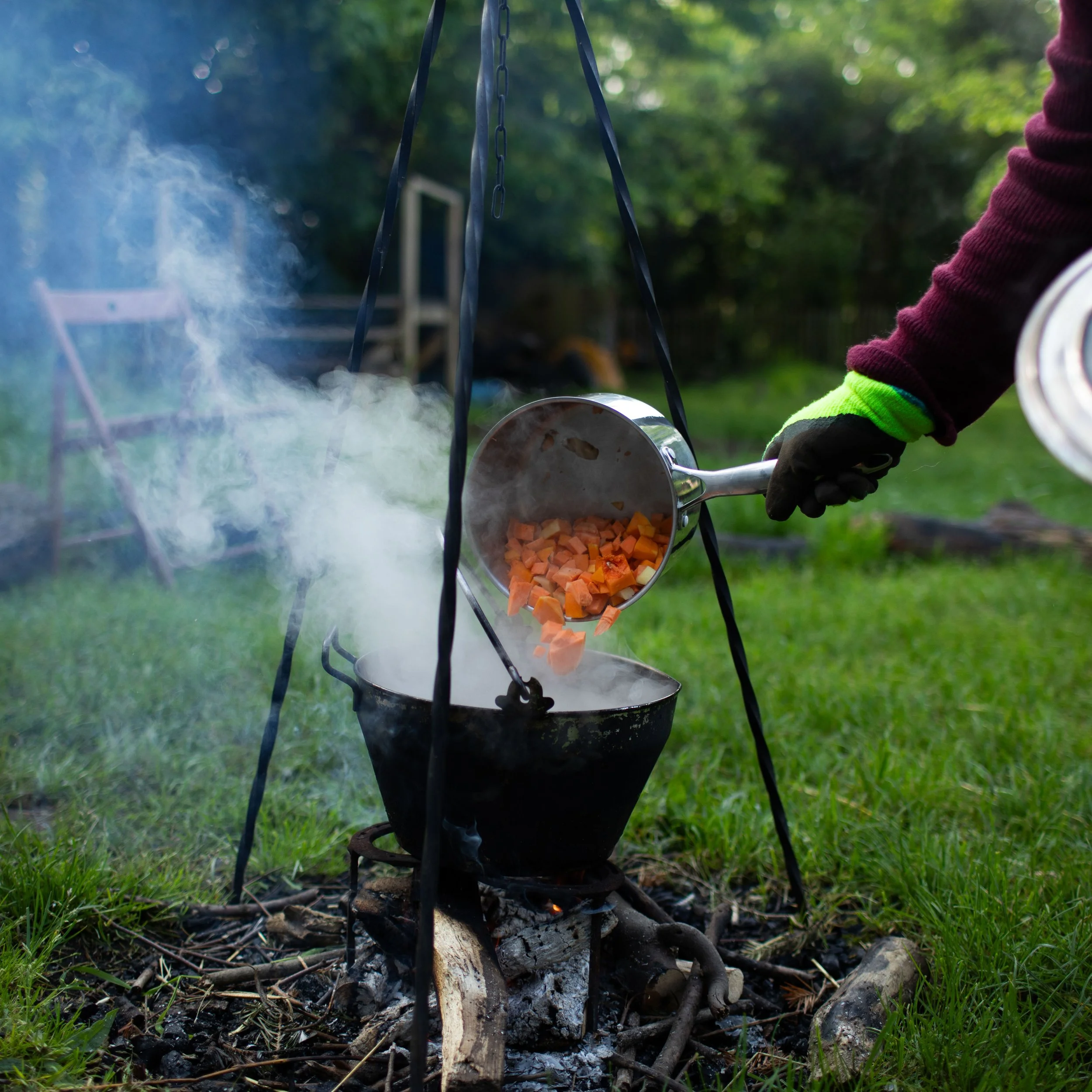 Person cooking chopped vegetables over an outdoor fire in a black pot, smoking visible, with a green grassy yard and trees in the background.