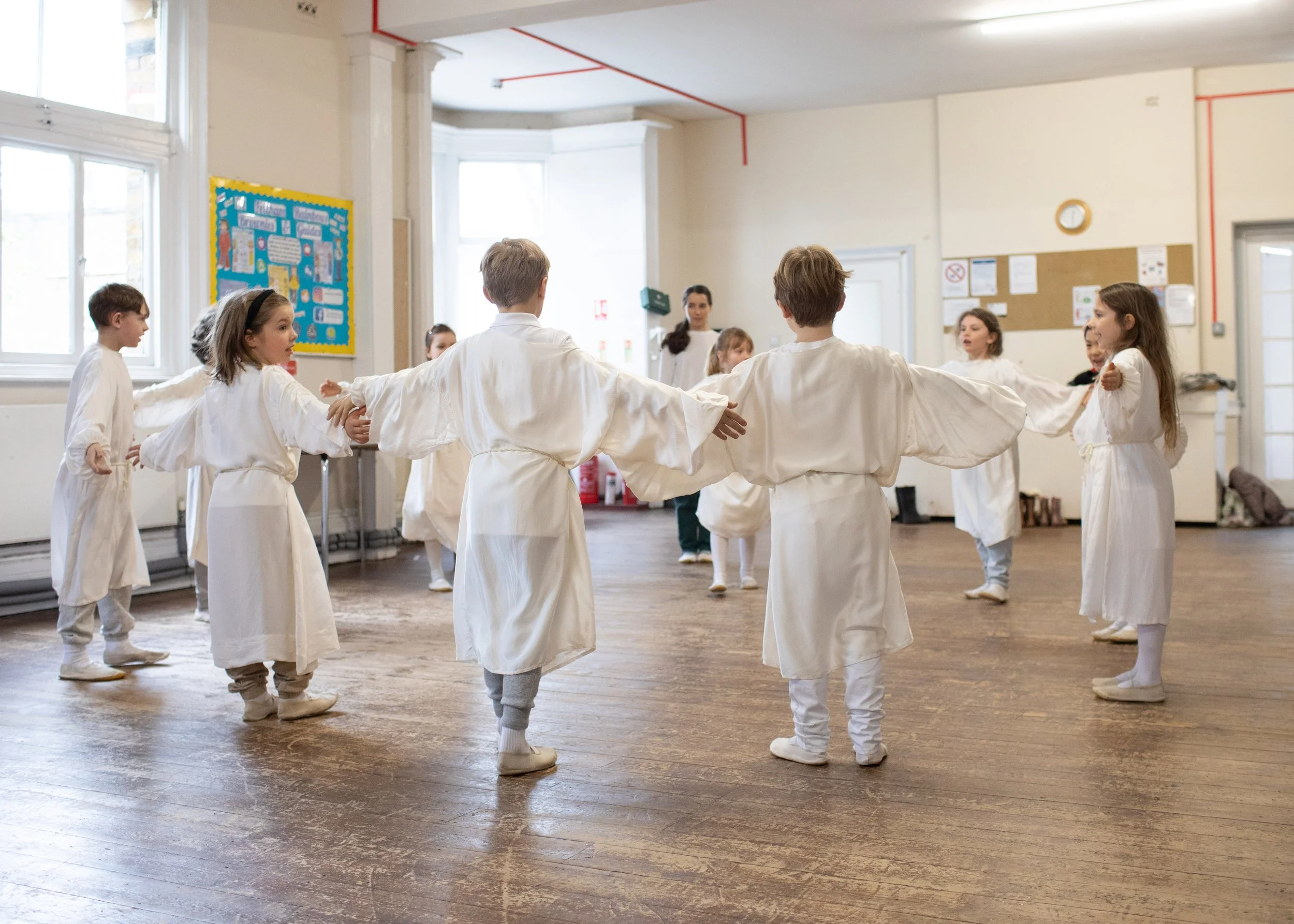 Children in white robes forming a circle and holding hands in a classroom or gymnasium.