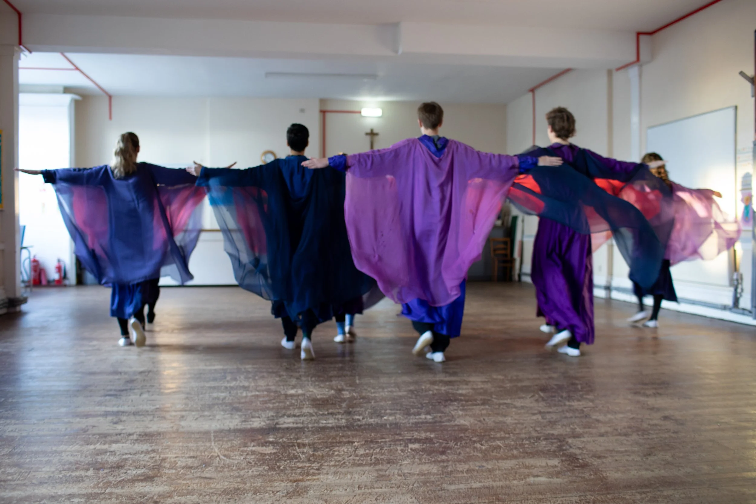 People in colorful flowing costumes practicing a dance in a room with wooden floor.