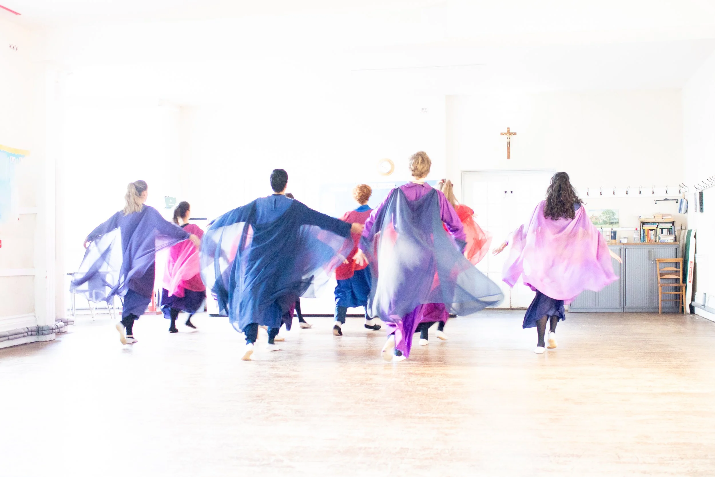 People practicing a dance or movement class in a bright room, wearing colorful flowing garments, with a cross on the wall and books on a shelf in the background.