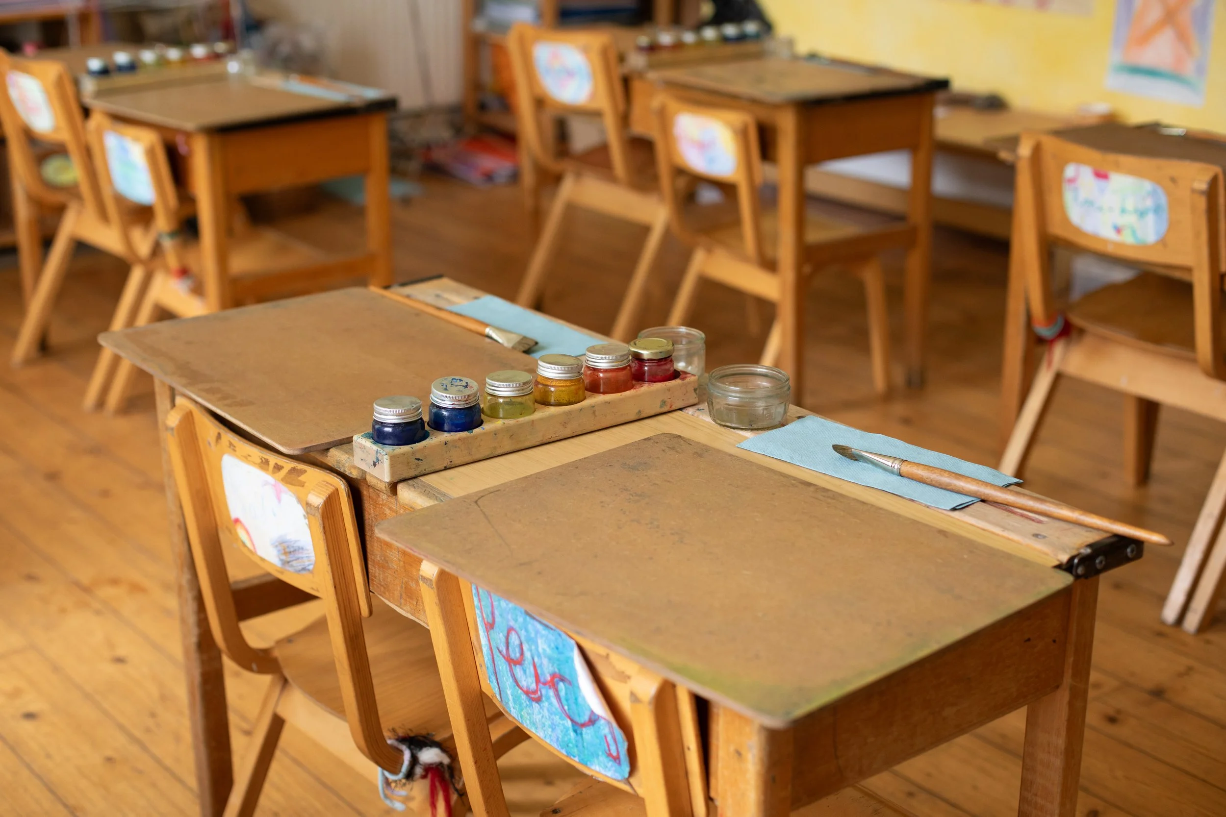 Child's art classroom with small wooden tables and chairs, art supplies including paints and brushes, and colorful drawings hanging on the walls.