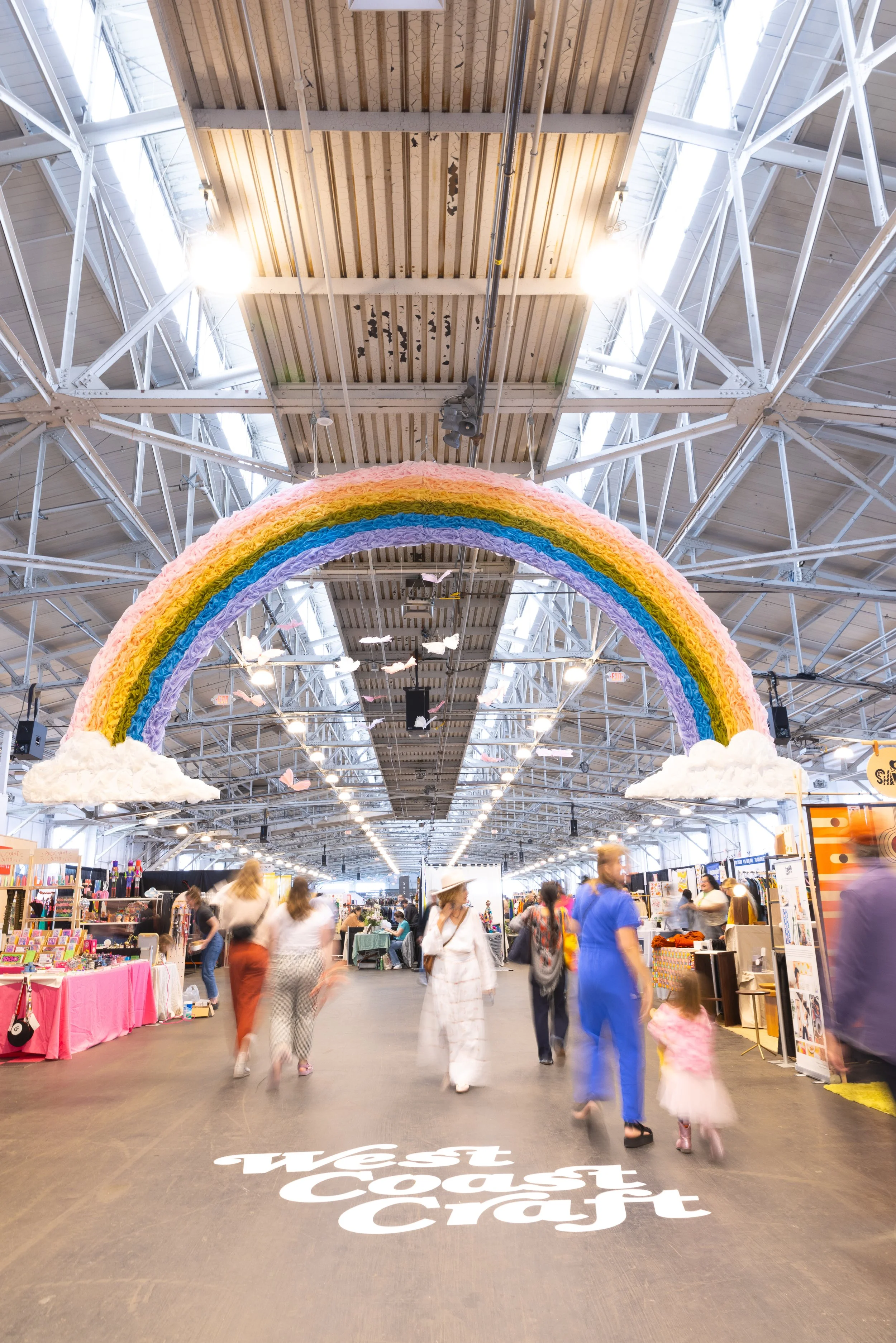 People walking through indoor market with oversized hanging rainbow decor.