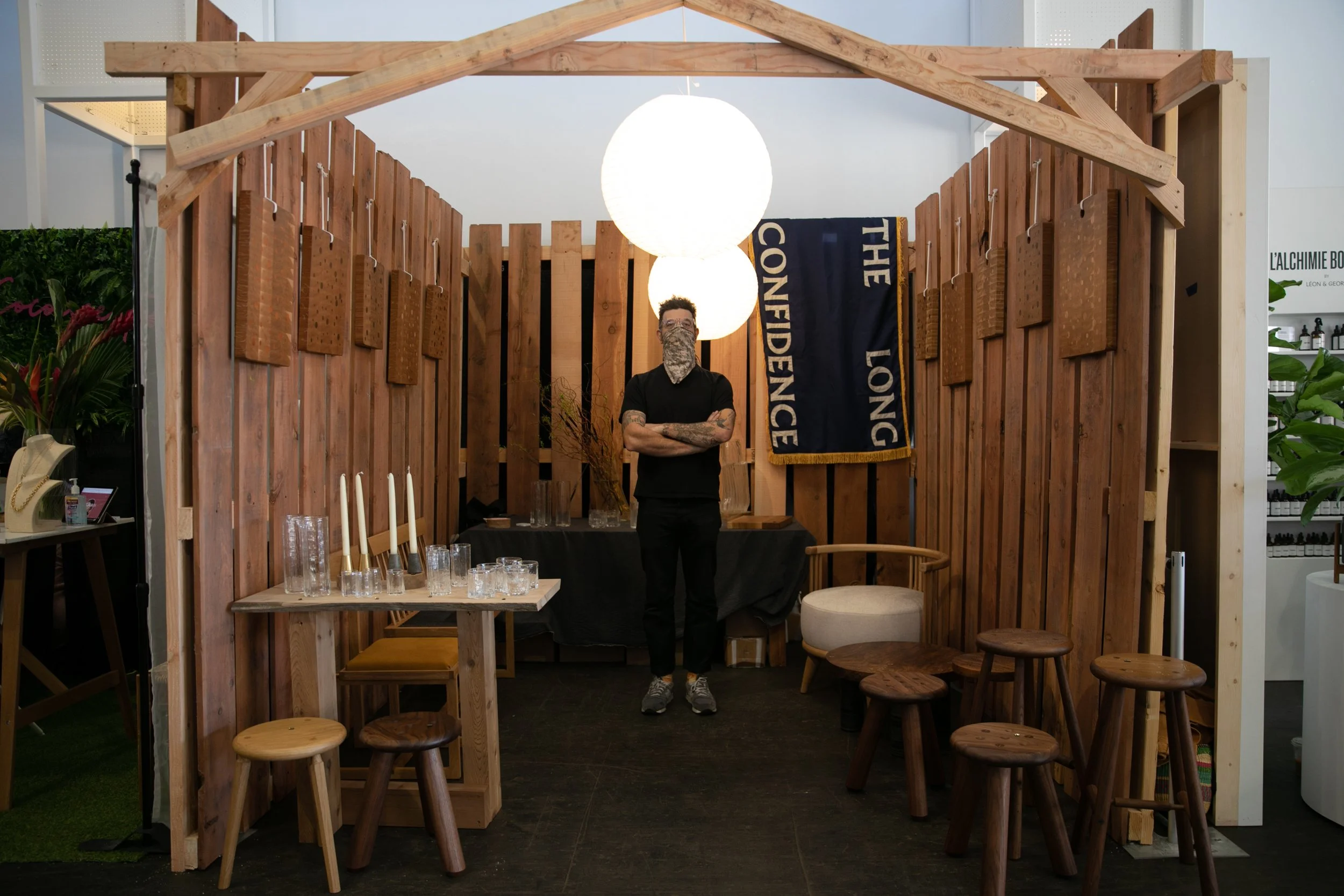 Man wearing a bandana mask standing in a wooden booth display with various home goods.