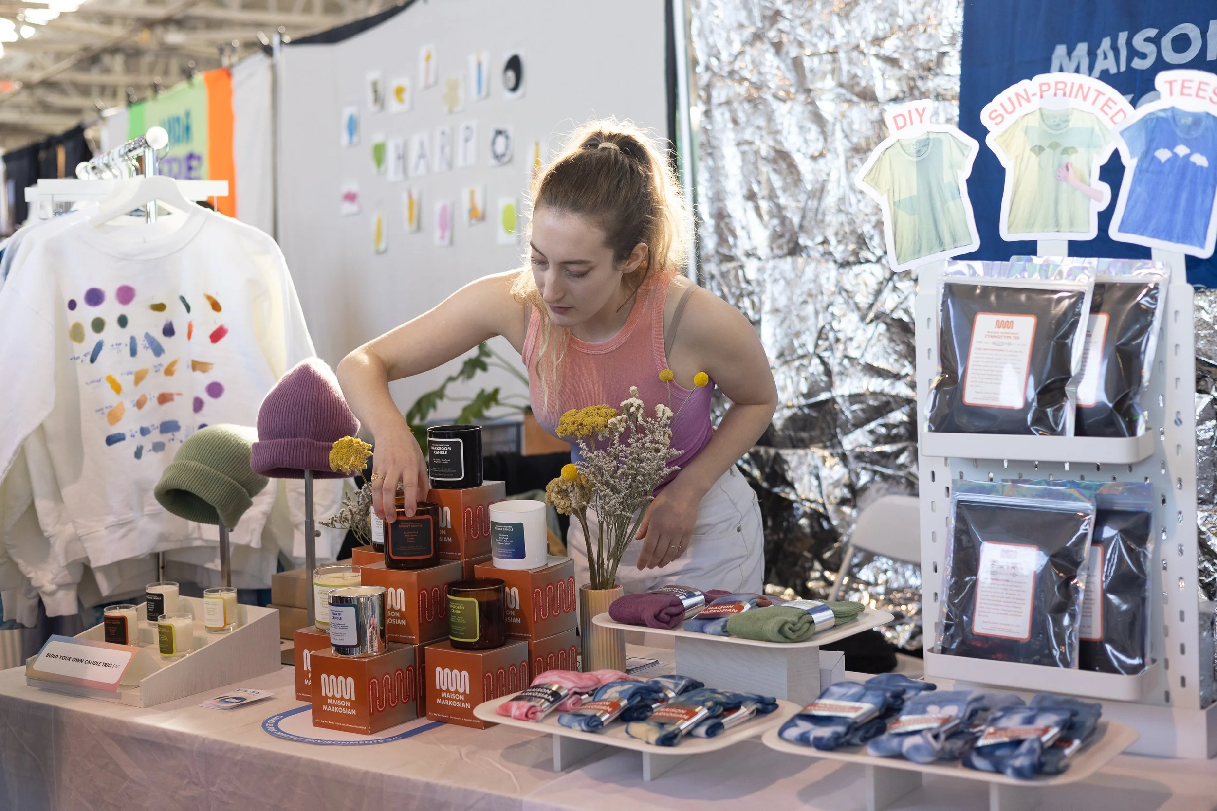 Woman adjusting candles stacked on a table displayed with various goods.