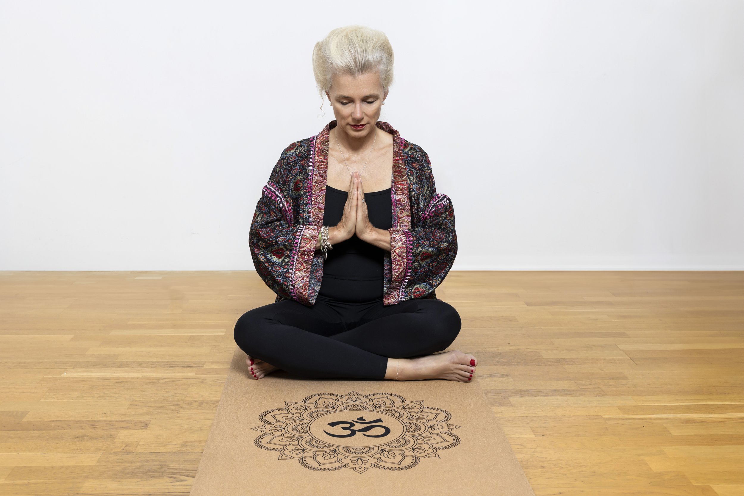 Frau in Yogapose sitzt im Schneidersitz auf einer Yogamatte mit Om-Symbol, trägt gemusterte Kleidung, Hände in Gebetshaltung, Holzfußboden.