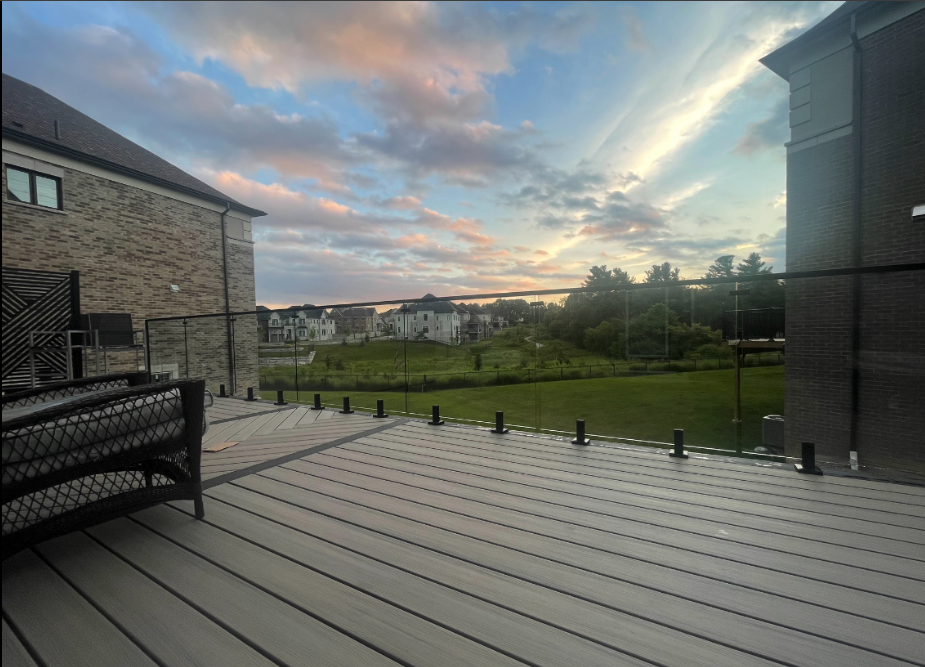 View of a balcony with a glass railing, outdoor furniture, and a green landscape with houses and trees in the distance during sunset.
