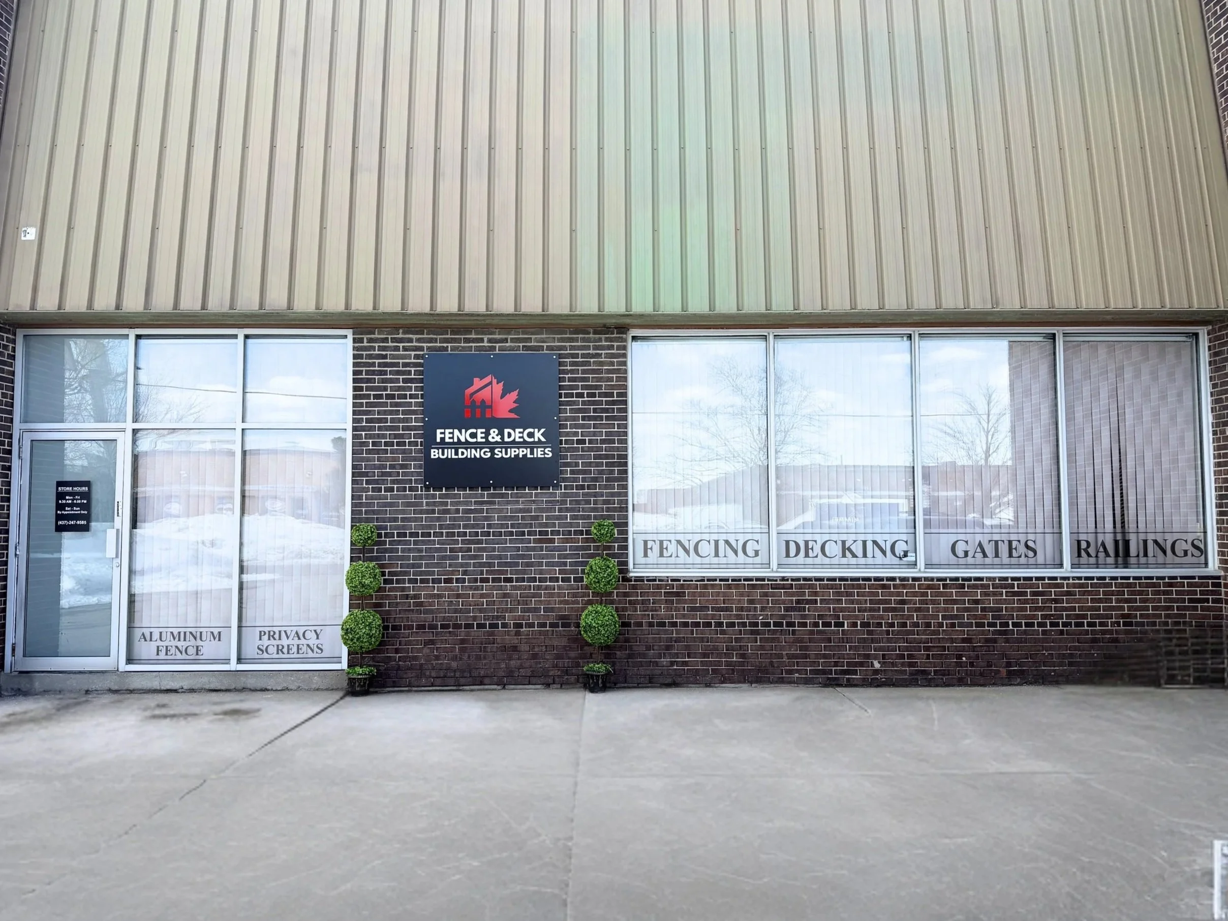  storefront with sign for Fence & Deck Building Supplies, brick and vinyl siding exterior, snow on ground, large windows advertising aluminum fence and privacy screens, reflections of trees and neighboring buildings in windows.