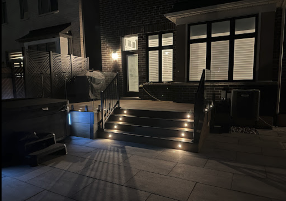 Night view of a backyard patio with stairs leading to a house door, illuminated by built-in step lights and a wall-mounted exterior light, with a hot tub and covered grill visible.