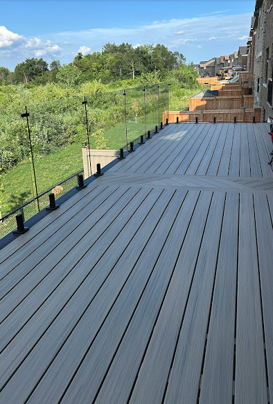 A spacious rooftop deck with gray composite decking, glass railing panels, and a view of green trees and partly cloudy sky in the background.