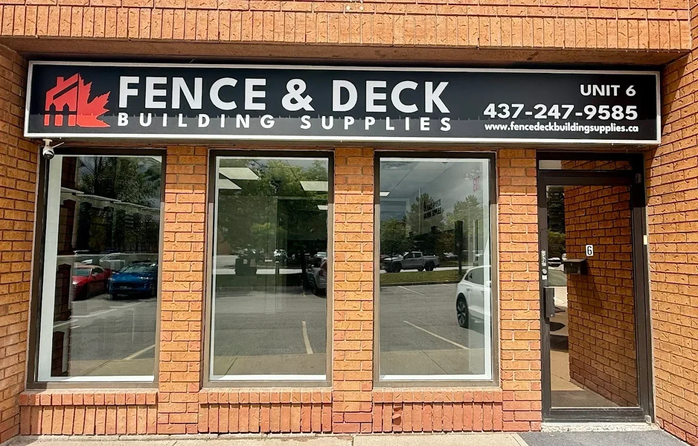 Storefront with a black sign reading 'FENCE & DECK Building Supplies' in white letters, brick exterior, large windows, and a glass door.