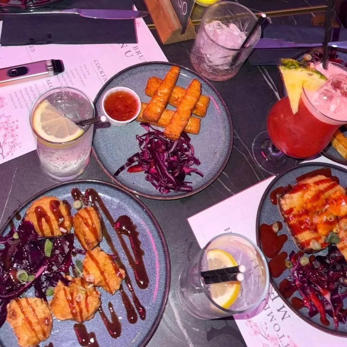 Dinner table with plates of fried food, purple cabbage salad, and drinks including lemon water and pink cocktails, with menus and cutlery visible.
