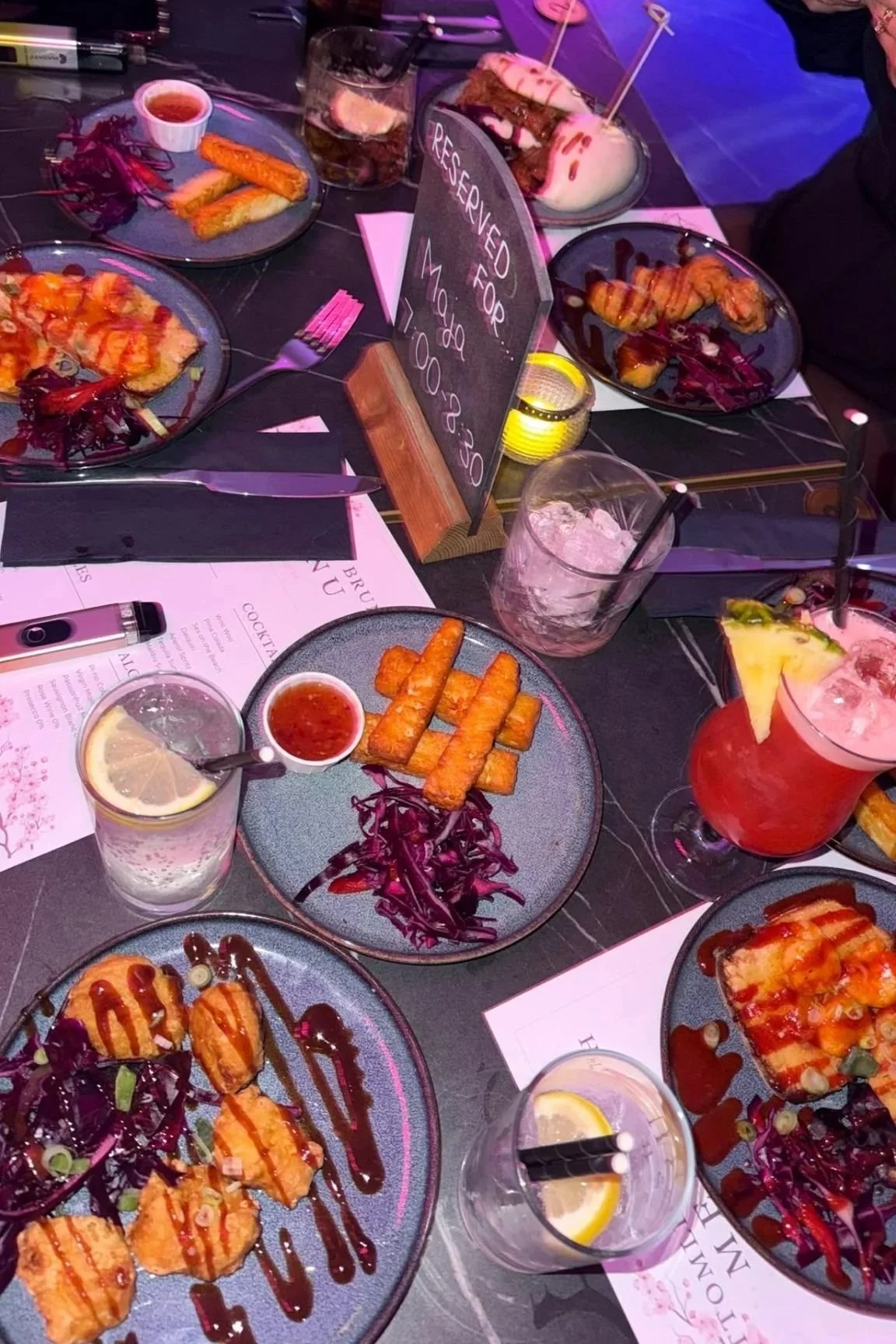 A table set with various plates of food, drinks, and a chalkboard sign that says, 'Reserved for, Mylo 6:30'. The plates contain food such as pizza, fried sticks with dip, grilled shrimp, and salad. There are drinks with lemon slices and slices of pineapple, including a cocktail with a cherry and a straw, a glass of water with lemon, and a cocktail with ice.