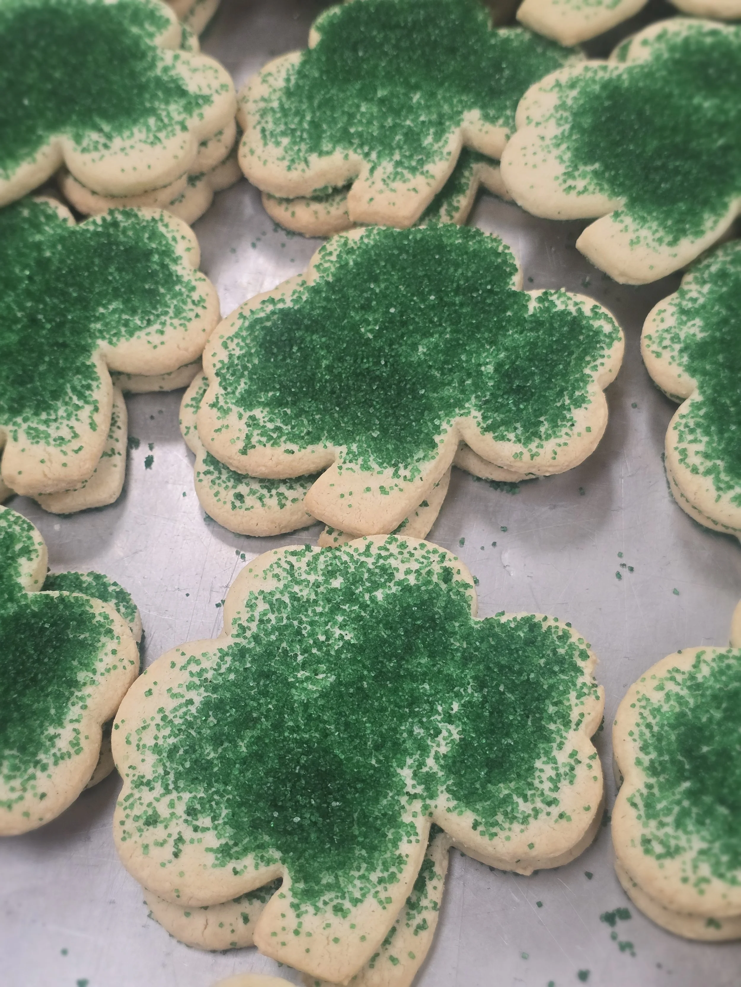 Cookies shaped like shamrocks with green sugar sprinkles on top, placed on a silver baking sheet.