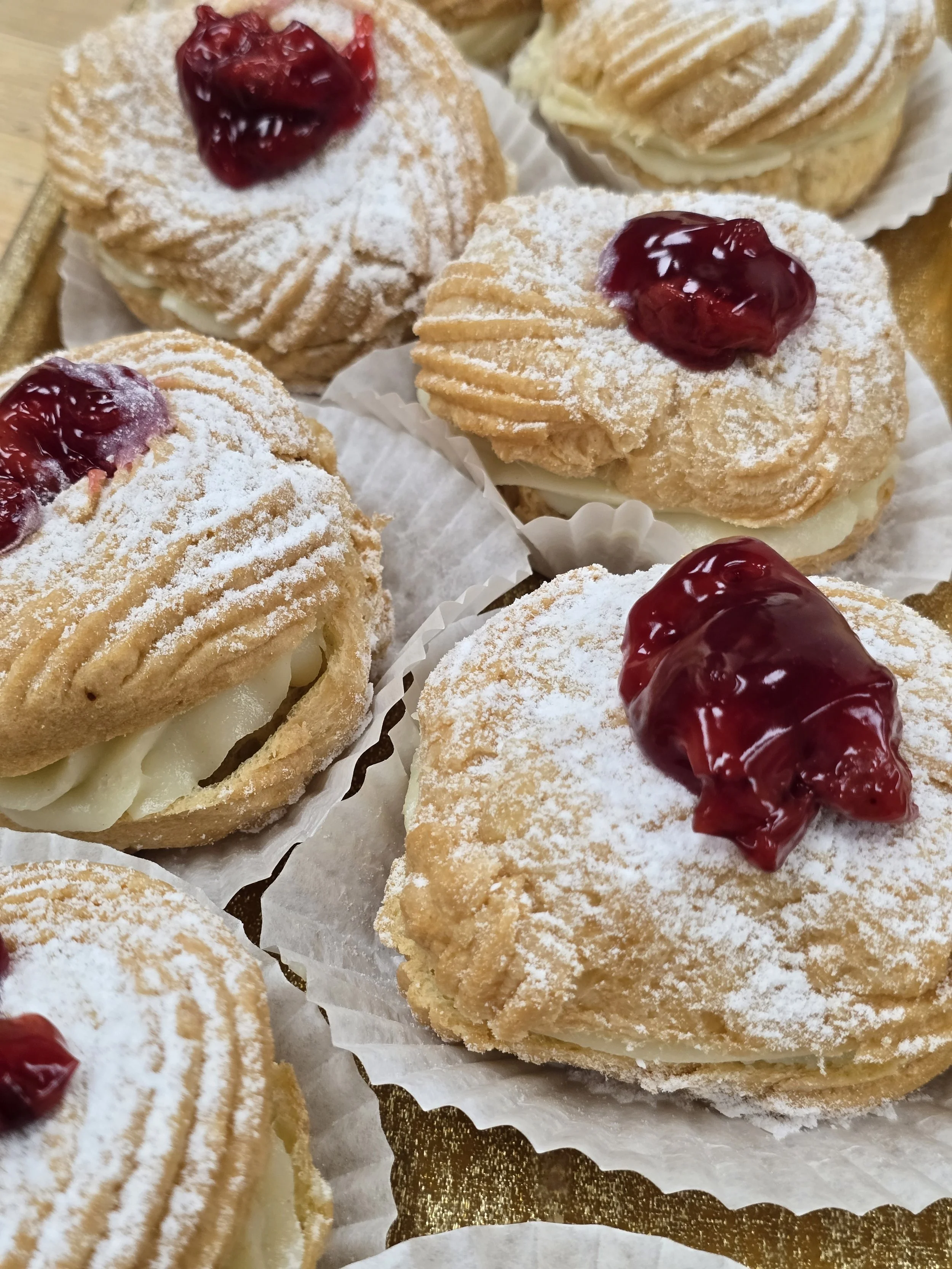 Cream-filled pastries topped with powdered sugar and cherry jam, arranged on a gold tray with paper cupcake liners.
