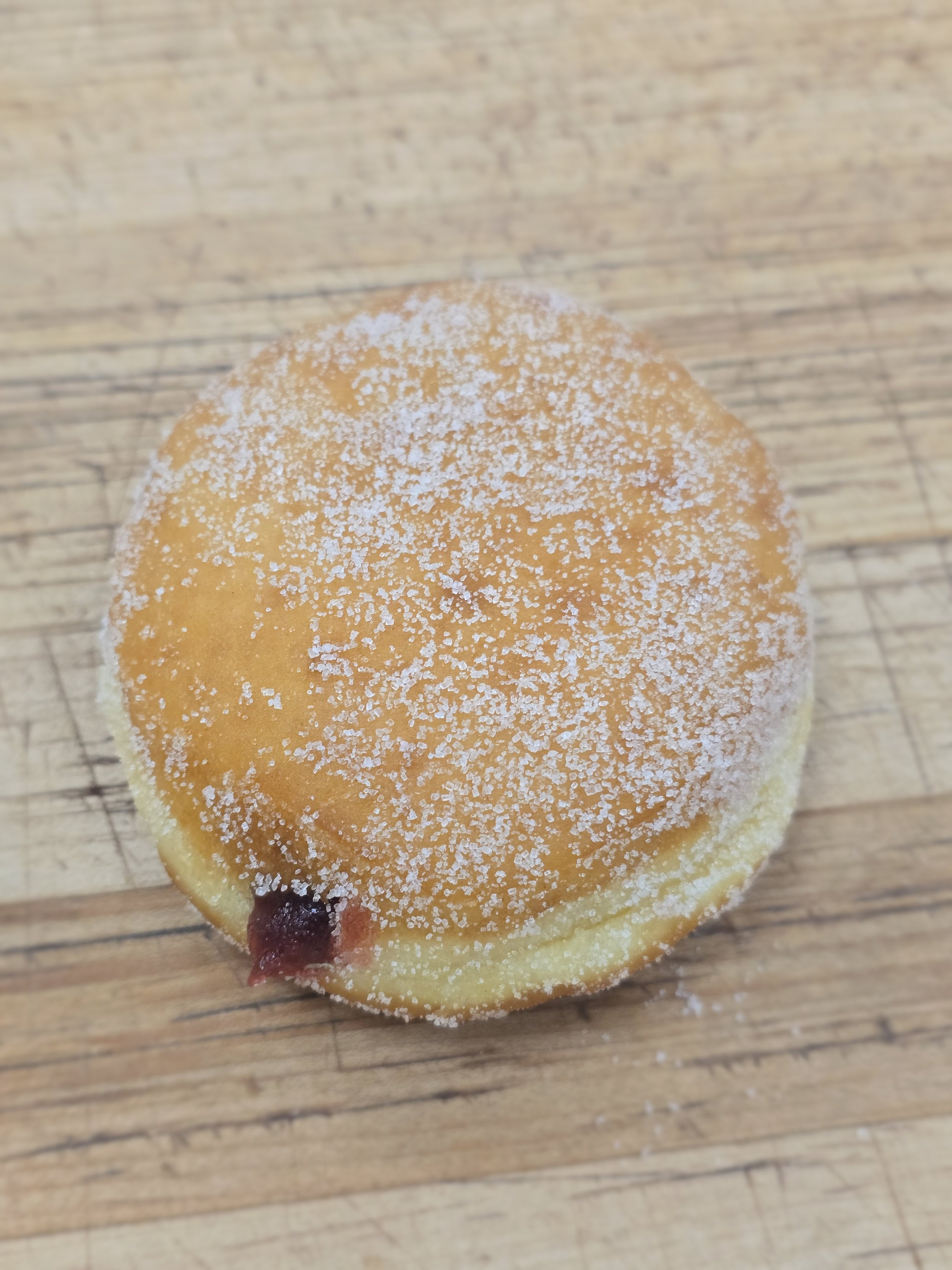 A round, sugar-dusted baked good with a cherry at the bottom edge, placed on a wooden surface.