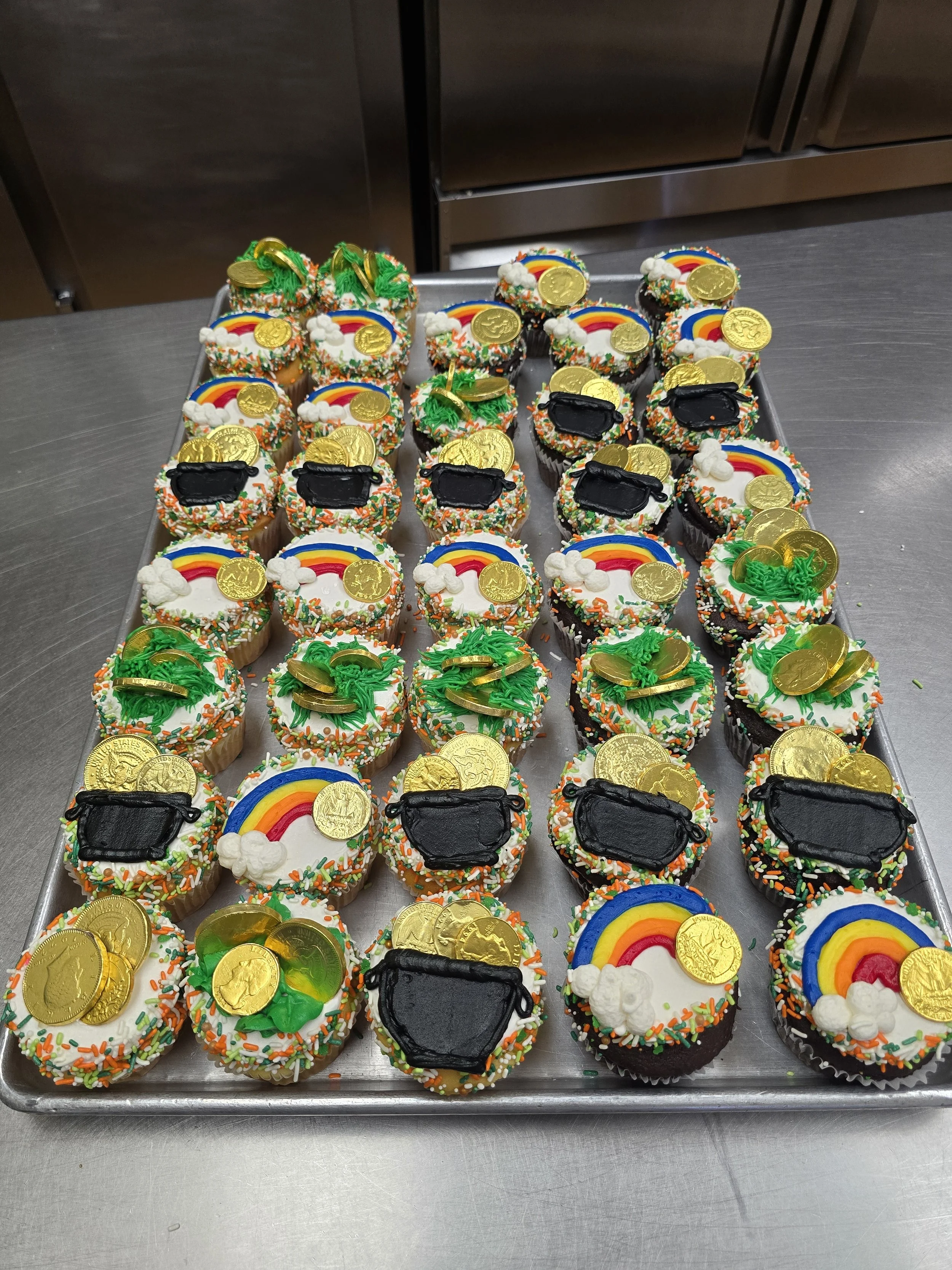A tray of decorated cupcakes with rainbow, gold coins, green money, and black face masks themed toppers, topped with colorful sprinkles.