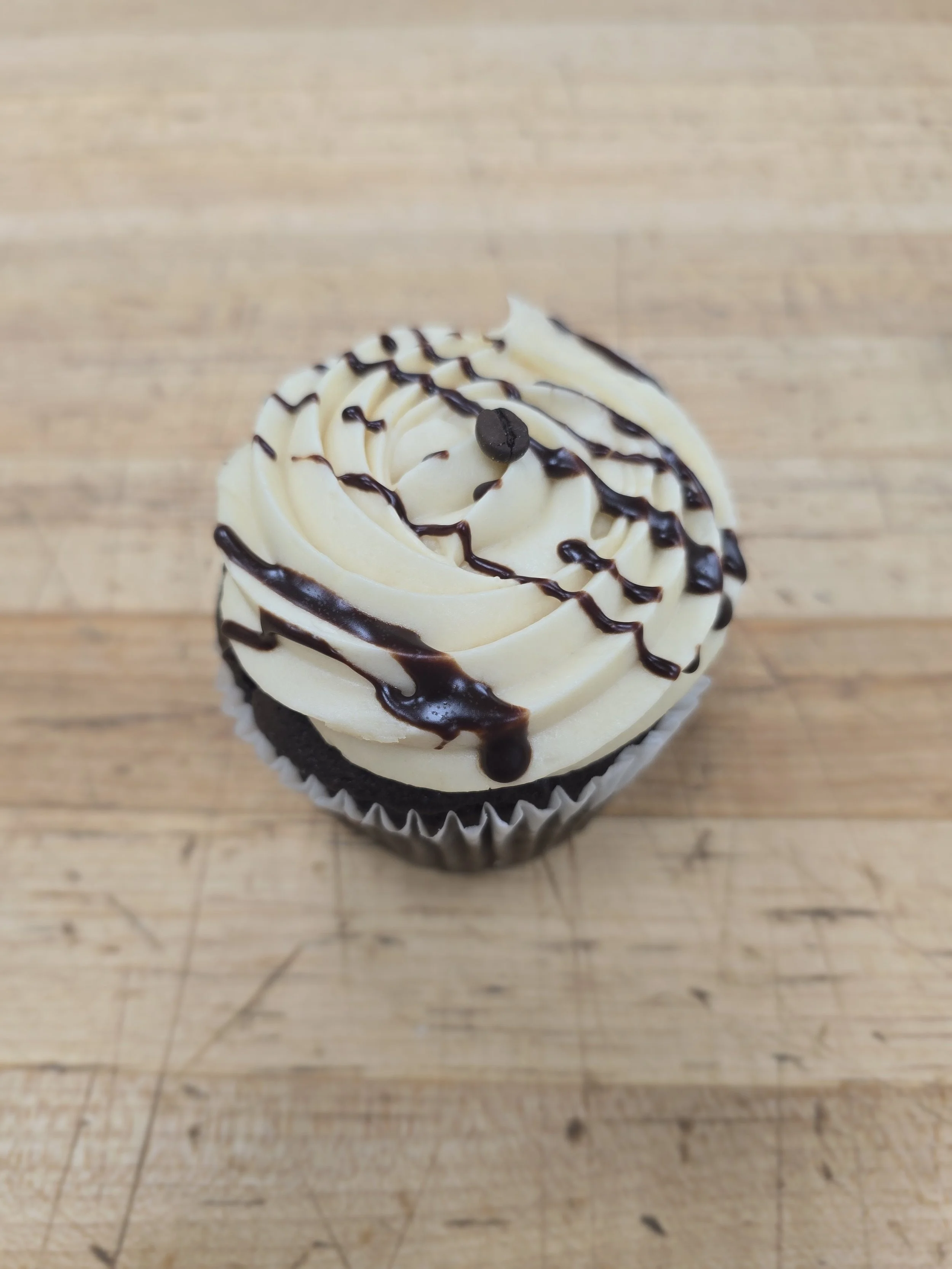 Chocolate cupcake with white frosting, drizzled with chocolate sauce, topped with a small chocolate piece, on a wooden surface.