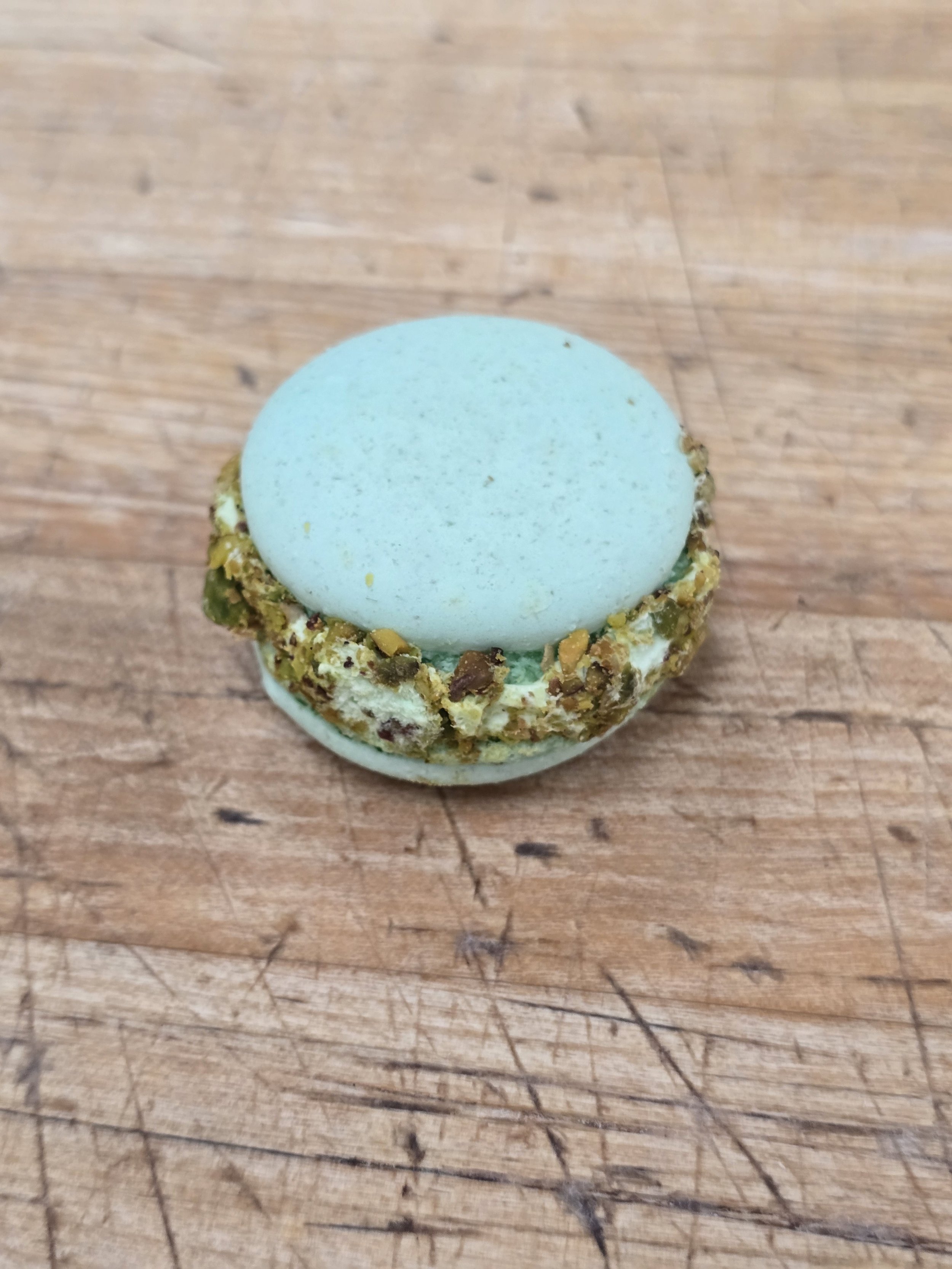 A light blue macaron on a wooden surface with a crunchy nut filling around the edges.