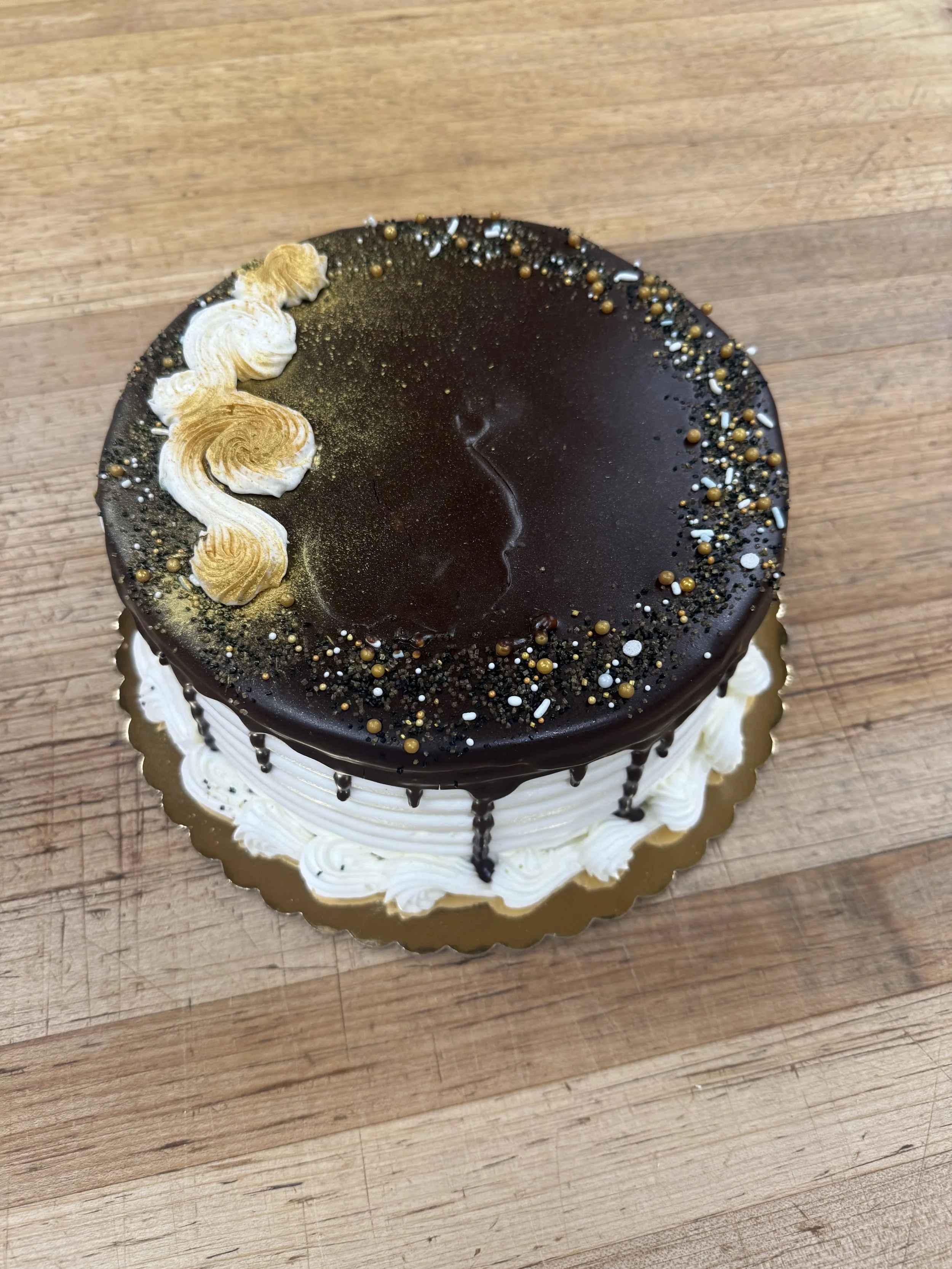 A round chocolate cake with white frosting, topped with chocolate shavings and a drip of chocolate on the sides. Garnished with chocolate curls and whipped cream on top.