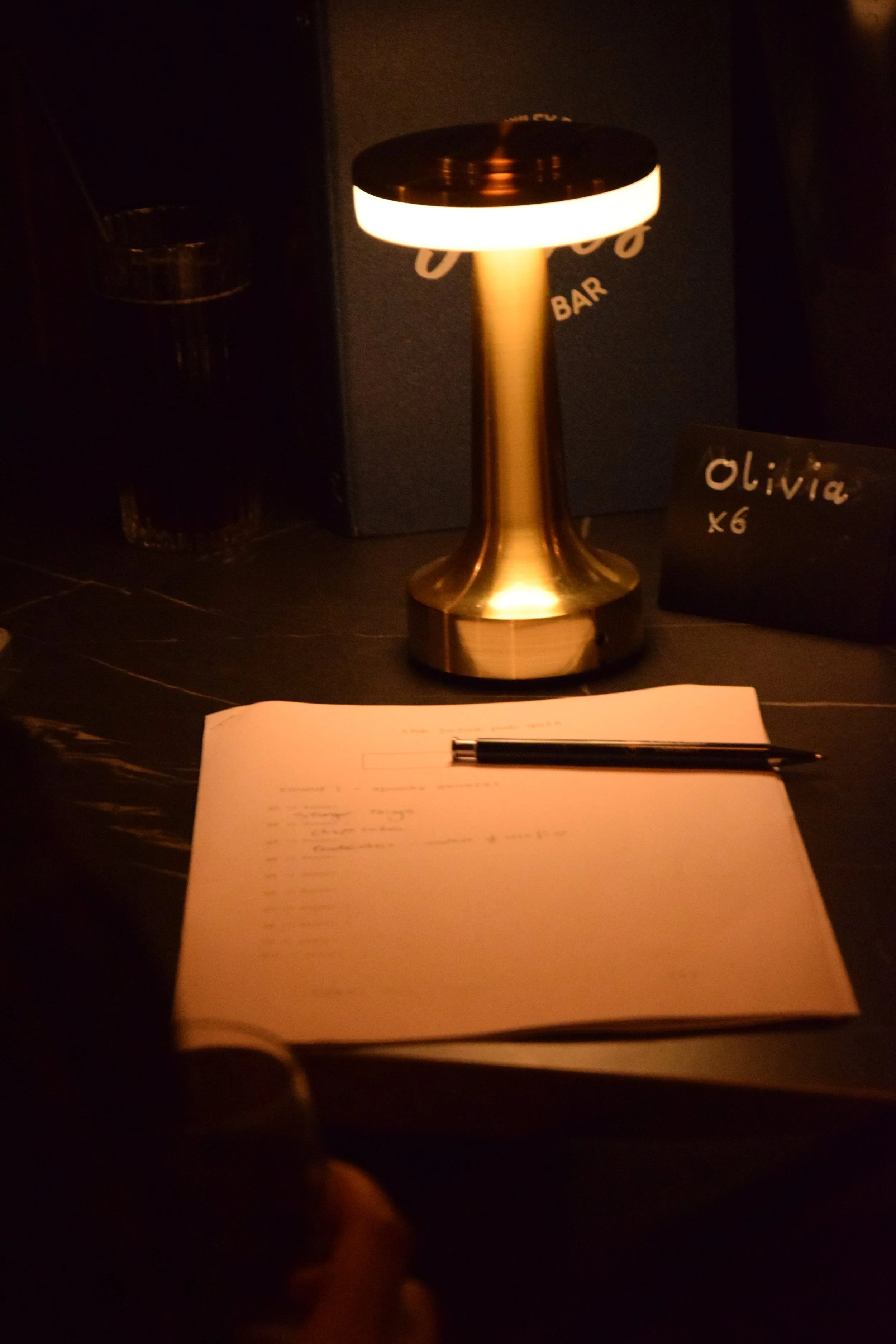 Dimly lit table with a sheet of paper and a pen for trivia, a brass lamp, and a small sign.