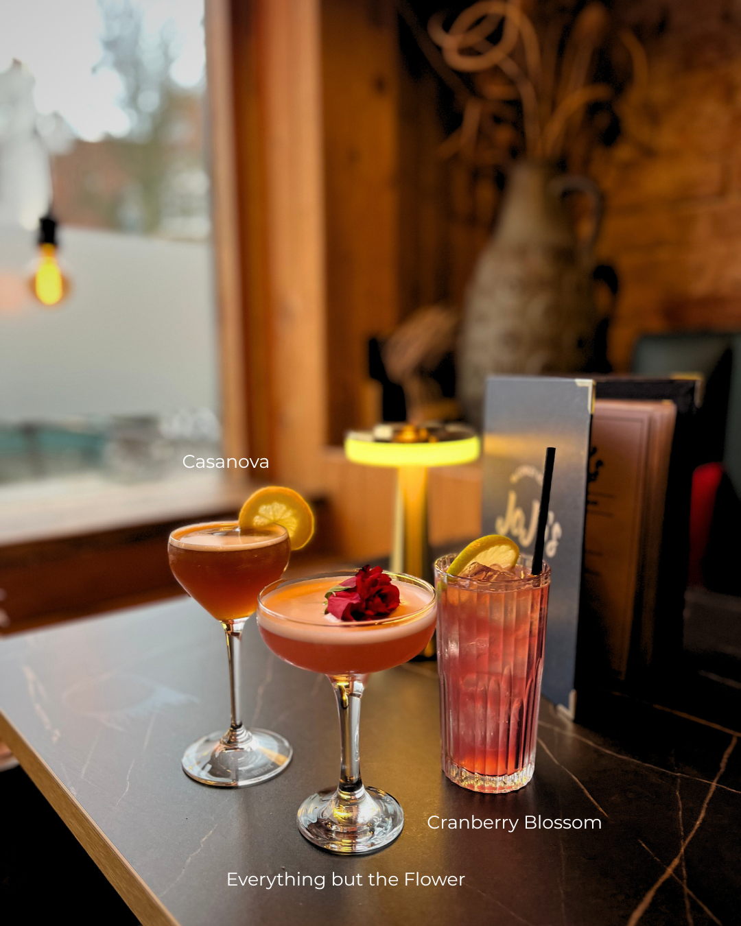 Three Valentine's Day cocktails on a table with a dimly lit, cozy background including a lamp, a large vase, and menus at JoJo's Cocktail & Pizza Bar, Cowley Road, Oxford.