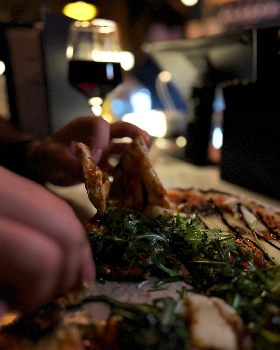 JoJo's customer reaching for a slice of pizza topped with greens with a glass of red wine in the background in a dimly lit cocktail bar in Cowley, Oxford.